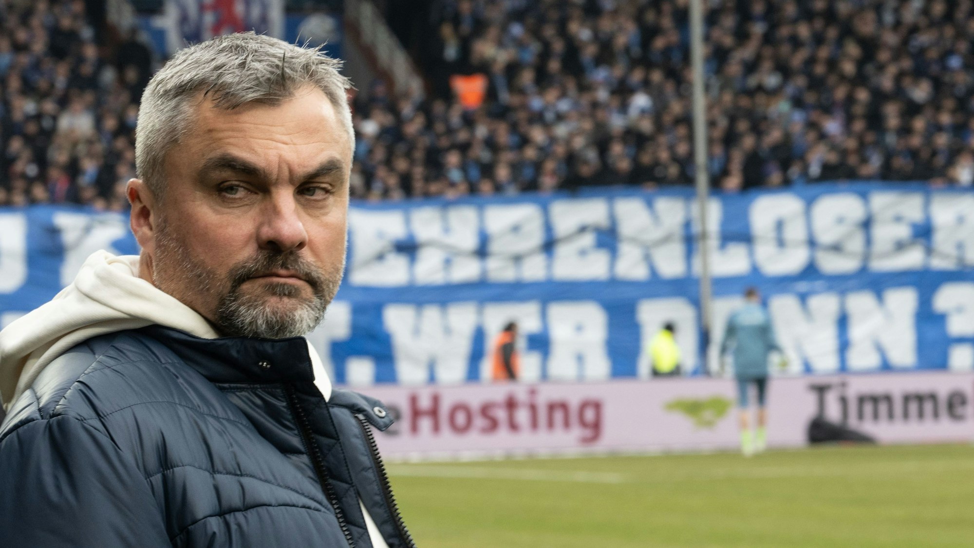Thomas Reis steht bei seiner Rückkehr ins Ruhrstadion des VfL Bochum am Spielfeldrand. Im Hintergrund hängt ein beleidigendes Plakat gegen den Trainer von Schalke 04.