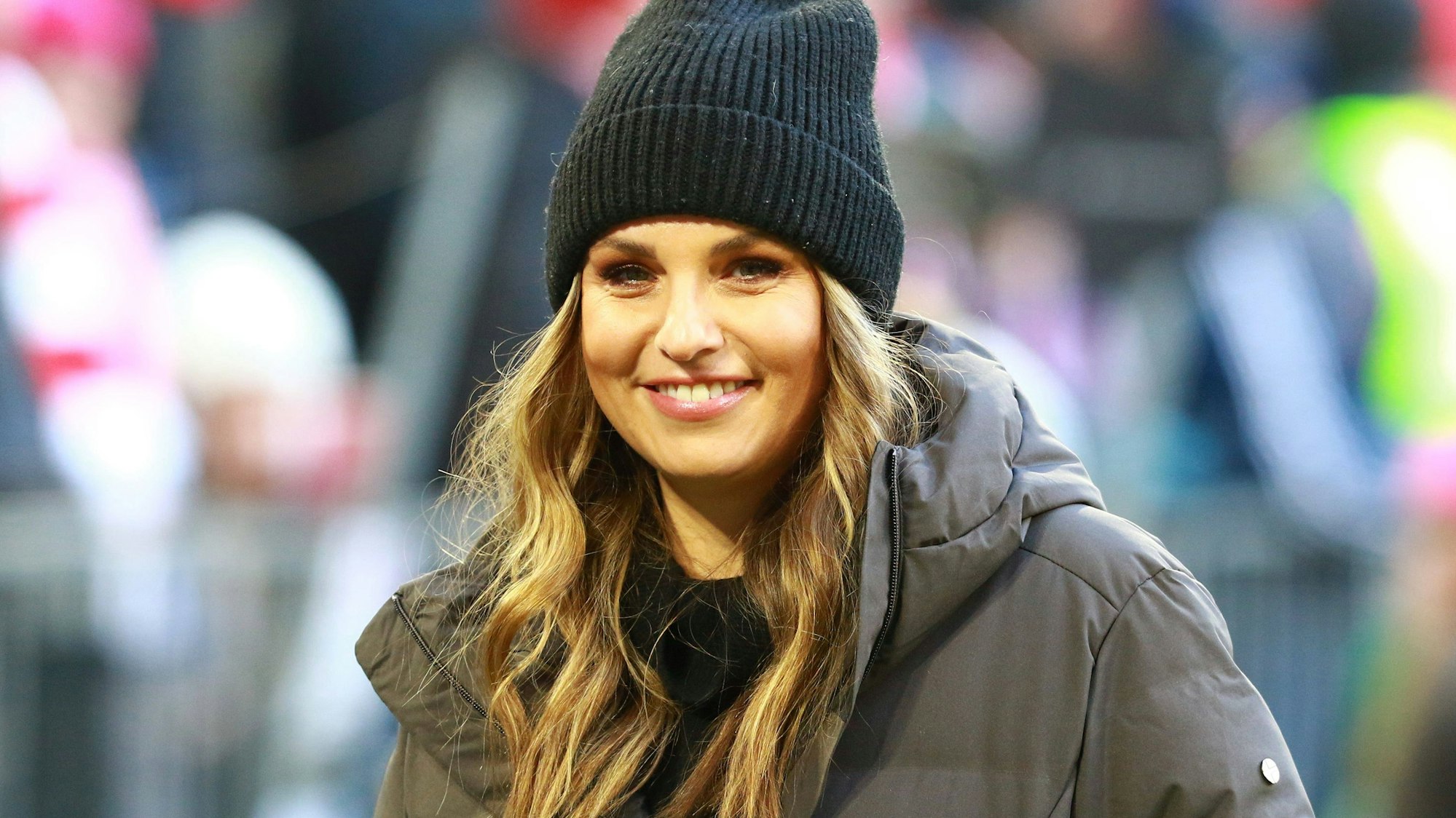 Laura Wontorra, mit Strickmütze bekleidet, lächelt vor dem Spiel Bayern München gegen Union Berlin in die Kamera.