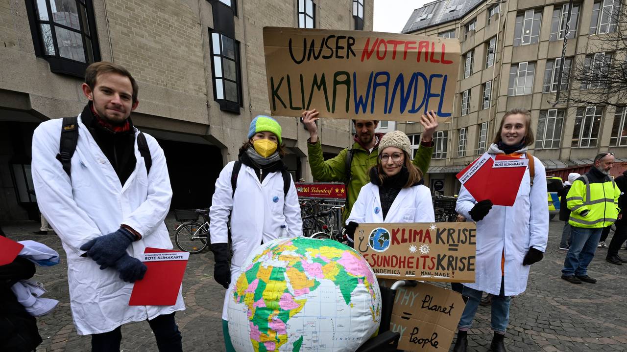 Demonstrierende von „Fridays for Future“ tragen weiße Kittel und halten beschriebene Plakate hoch. Auf einem Rollstuhl vor ihnen liegt eine Weltkugel.