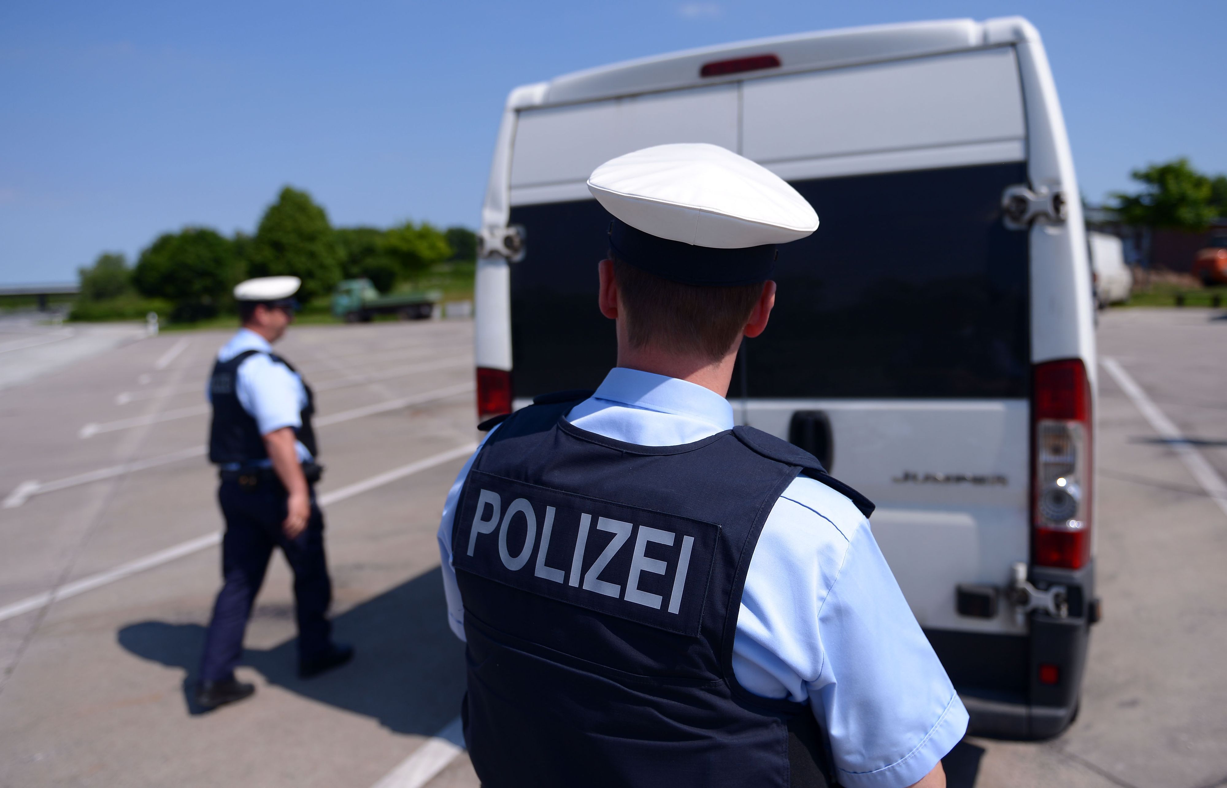 Das Symbolfoto zeigt zwei deutsche Polizisten in Uniform. Sie stehen vor einem weißen Transporter auf einem leeren Parkplatz.