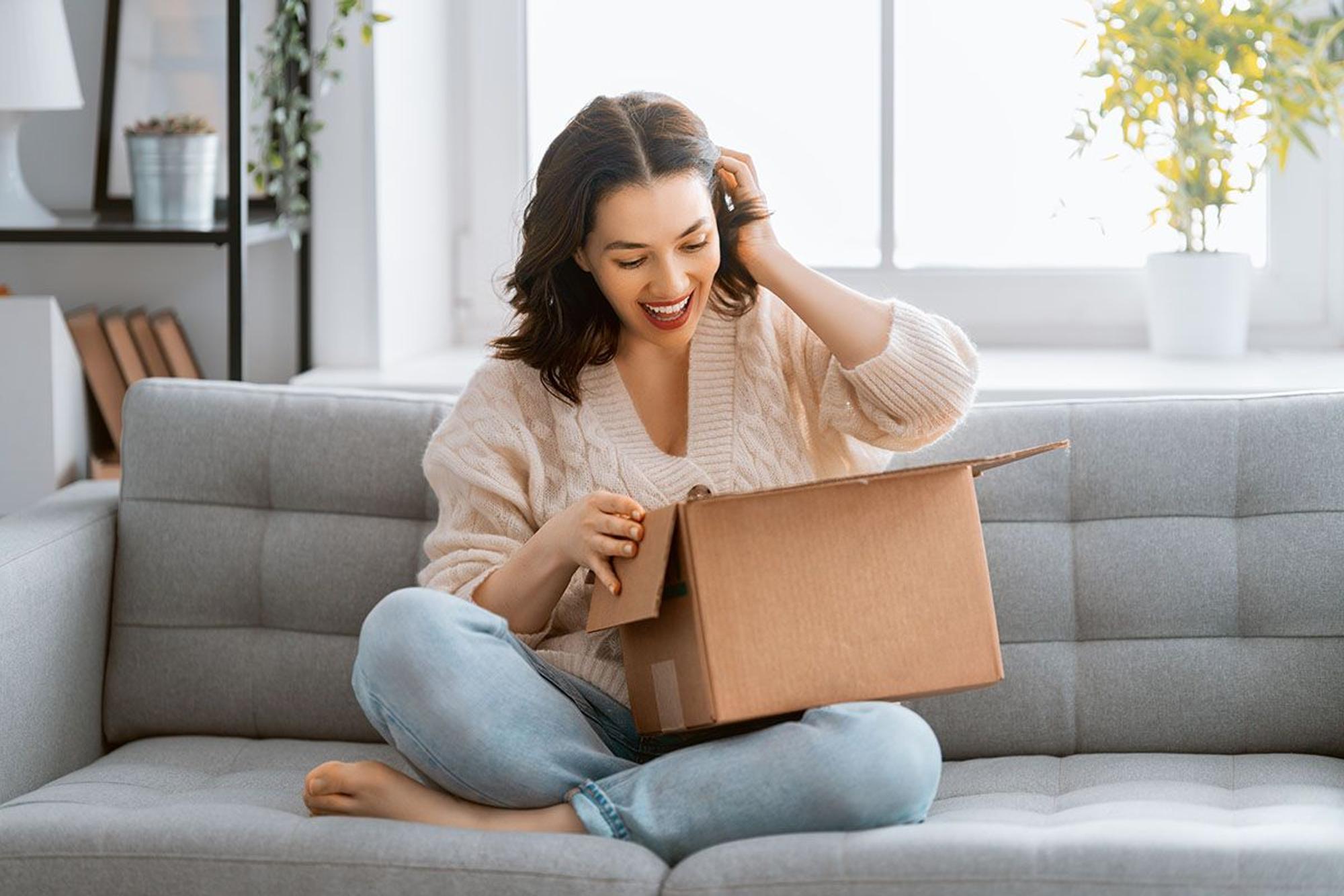 Junge Frau sitzt auf der Couch und packt freudig ein Paket aus