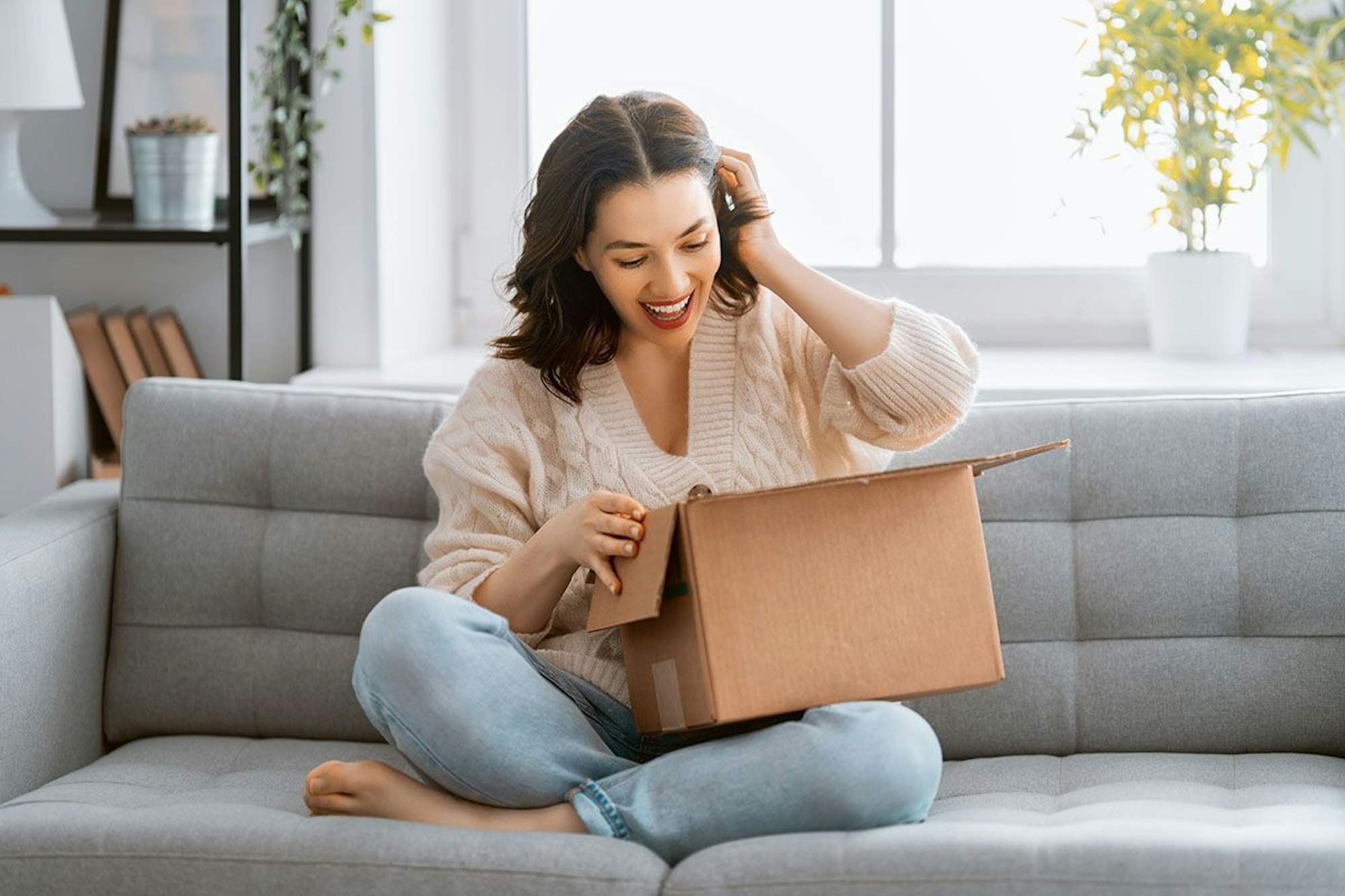 Junge Frau sitzt auf der Couch und packt freudig ein Paket aus