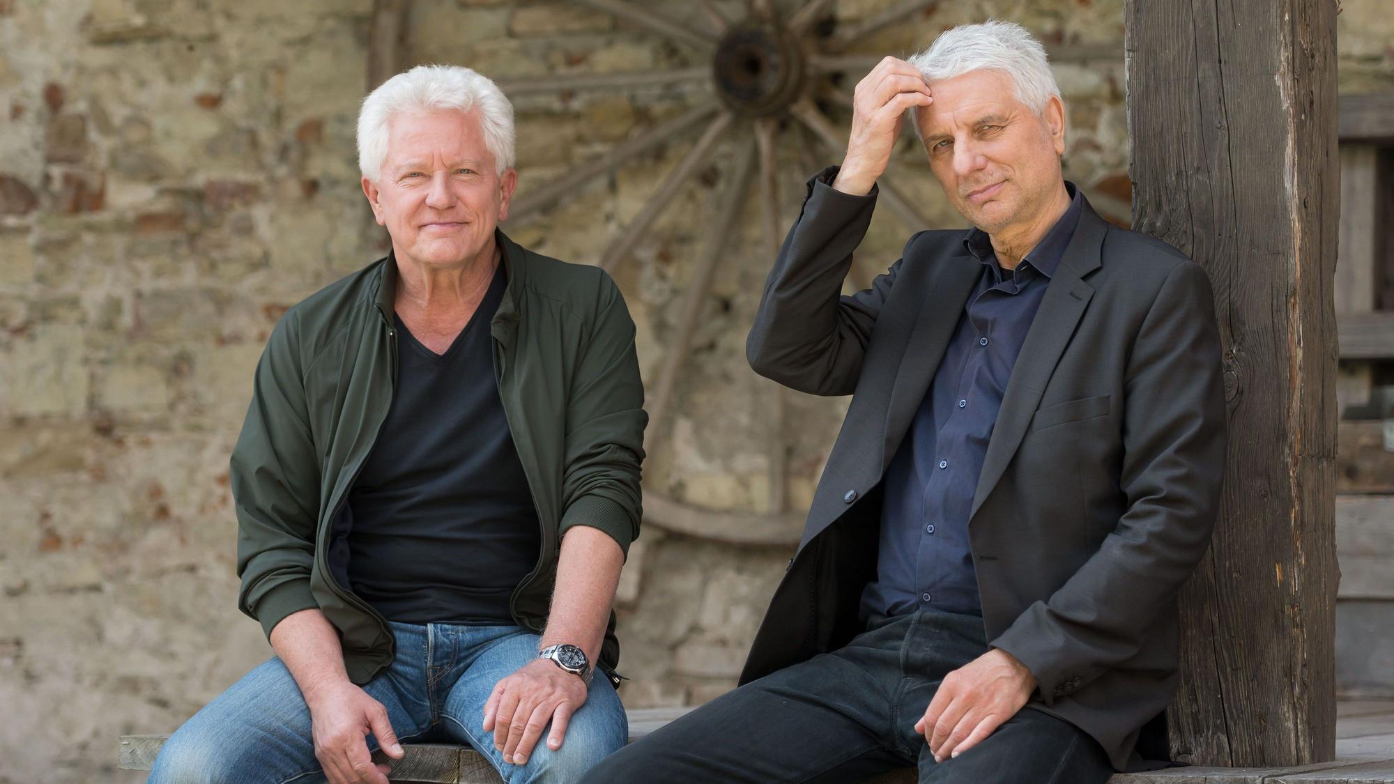Die Schauspieler Miroslav Nemec und Udo Wachtveitl posieren für ein Foto. Beide Schauspieler sitzen auf einer Holzbank.