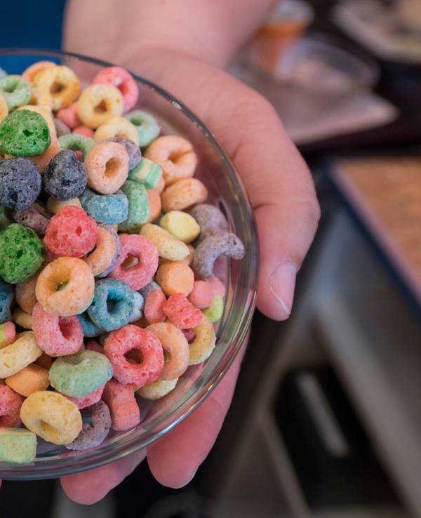 Das Symbolfoto aus dem Jahr 2018 zeigt eine Glasschüssel mit bunten, zu Ringen geformten Cornflakes. Die Schüssel wird von zwei Händen gehalten.