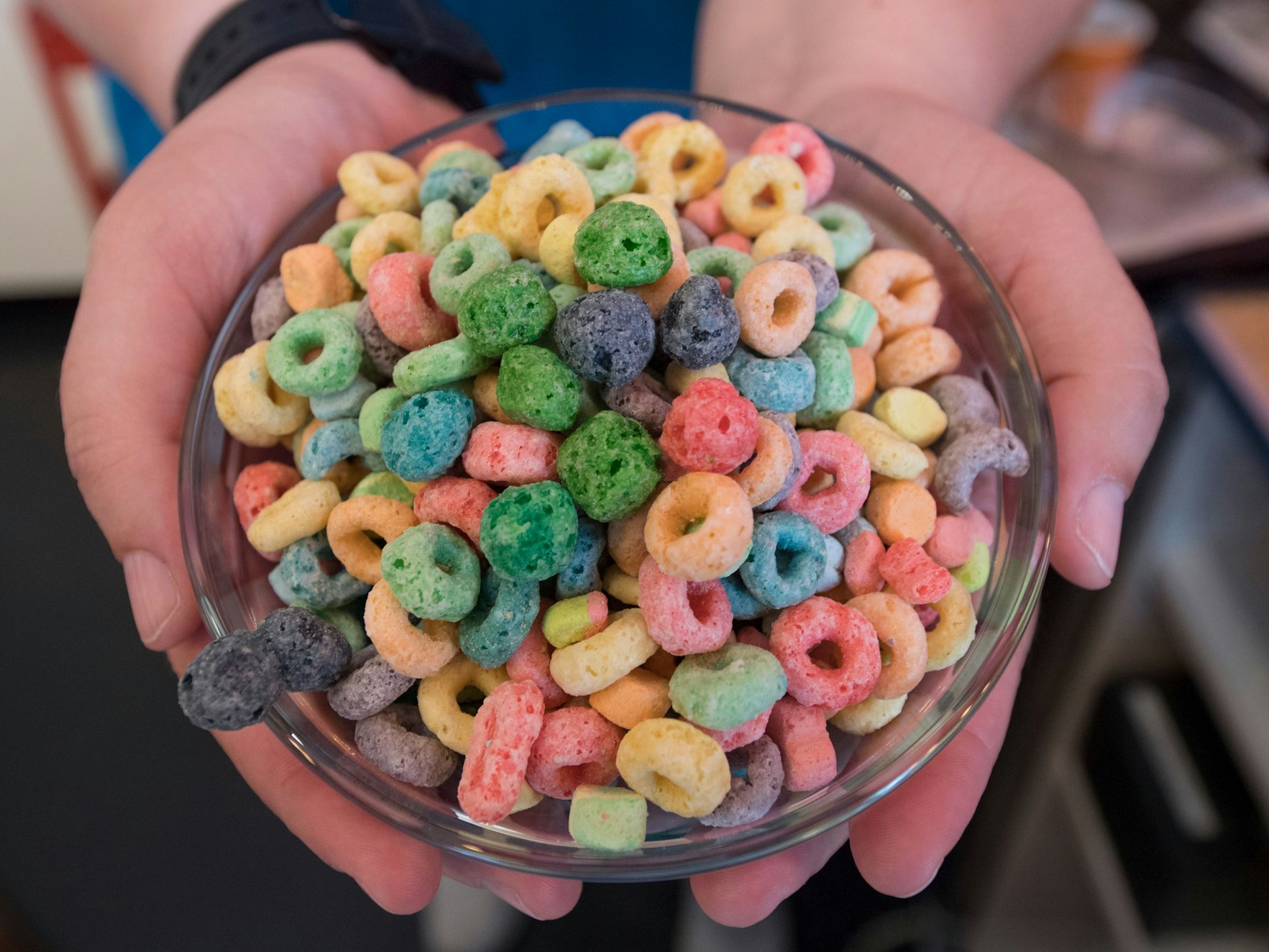 Das Symbolfoto aus dem Jahr 2018 zeigt eine Glasschüssel mit bunten, zu Ringen geformten Cornflakes. Die Schüssel wird von zwei Händen gehalten.