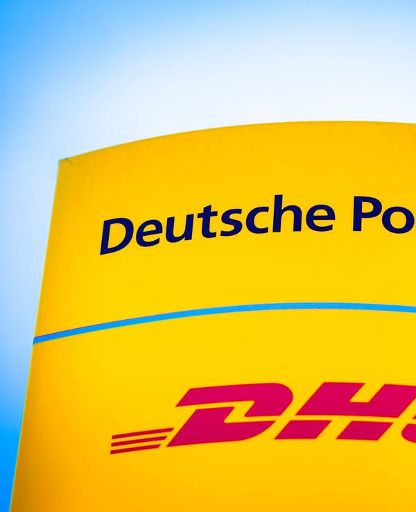 Die Logos von Deutsche Post und DHL leuchten am Morgen auf einem Schild vor einer Zustellbasis.