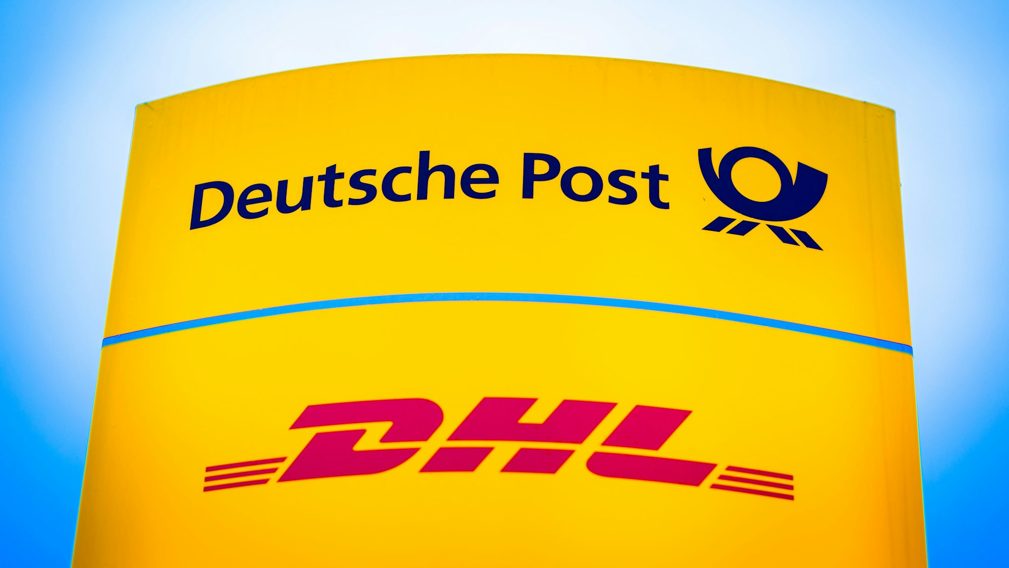Die Logos von Deutsche Post und DHL leuchten am Morgen auf einem Schild vor einer Zustellbasis.