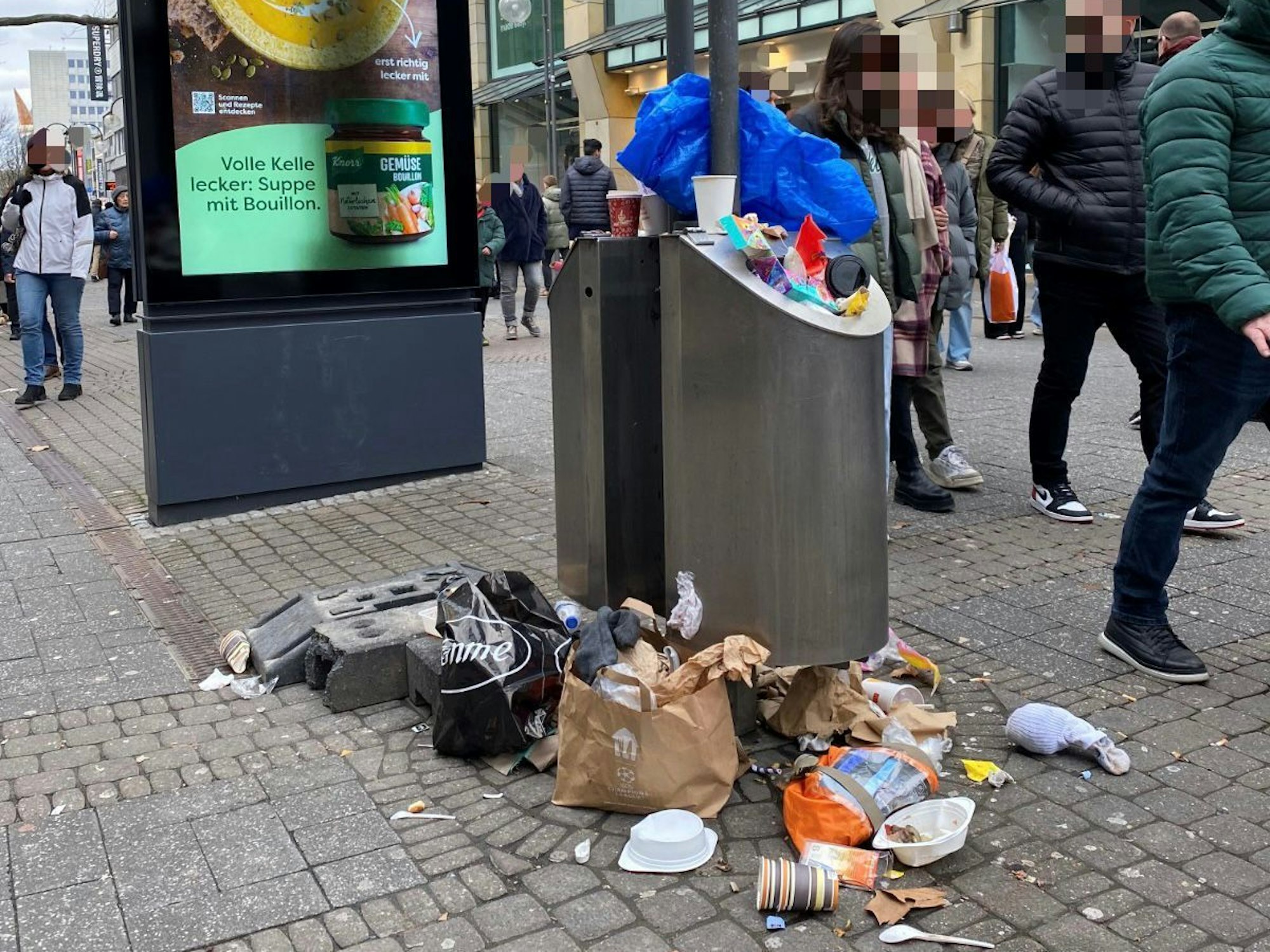 Menschen laufen an einem Mülleimer vorbei, der überquillt. Müll liegt bereits auf dem Boden.
