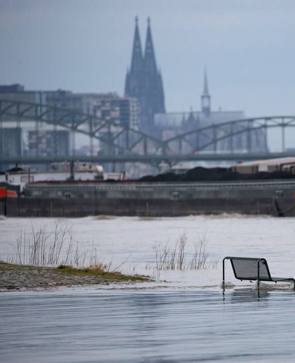 Binnenschiffe fahren auf dem Rhein vor der Kulisse des Kölner Doms, im Vordergrund steht eine vom Wasser umspülte Sitzbank. Noch hat der Kölner Pegel die Hochwassermarke nicht überschritten.