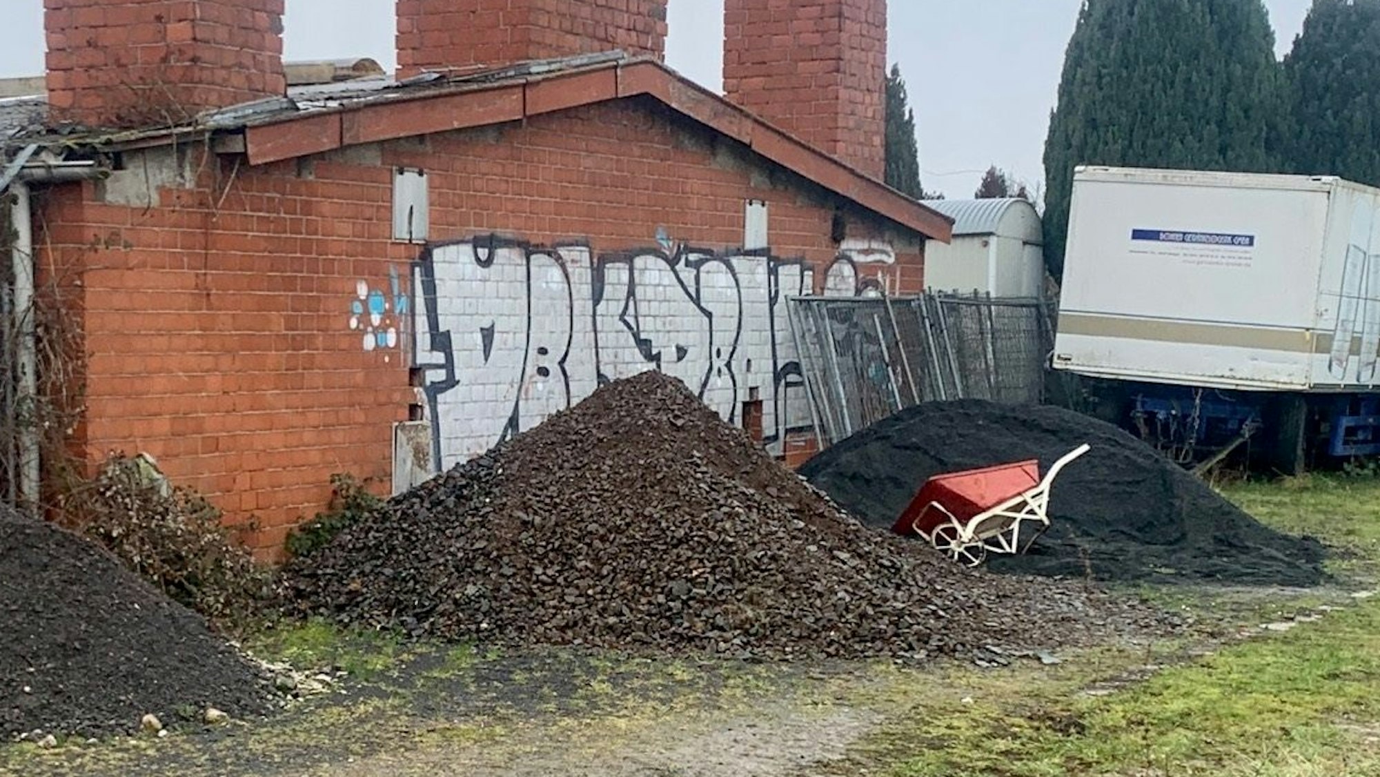 Mehrere Steinhaufen liegen vor einem Gebäude, an einem lehnt eine Schubkarre.