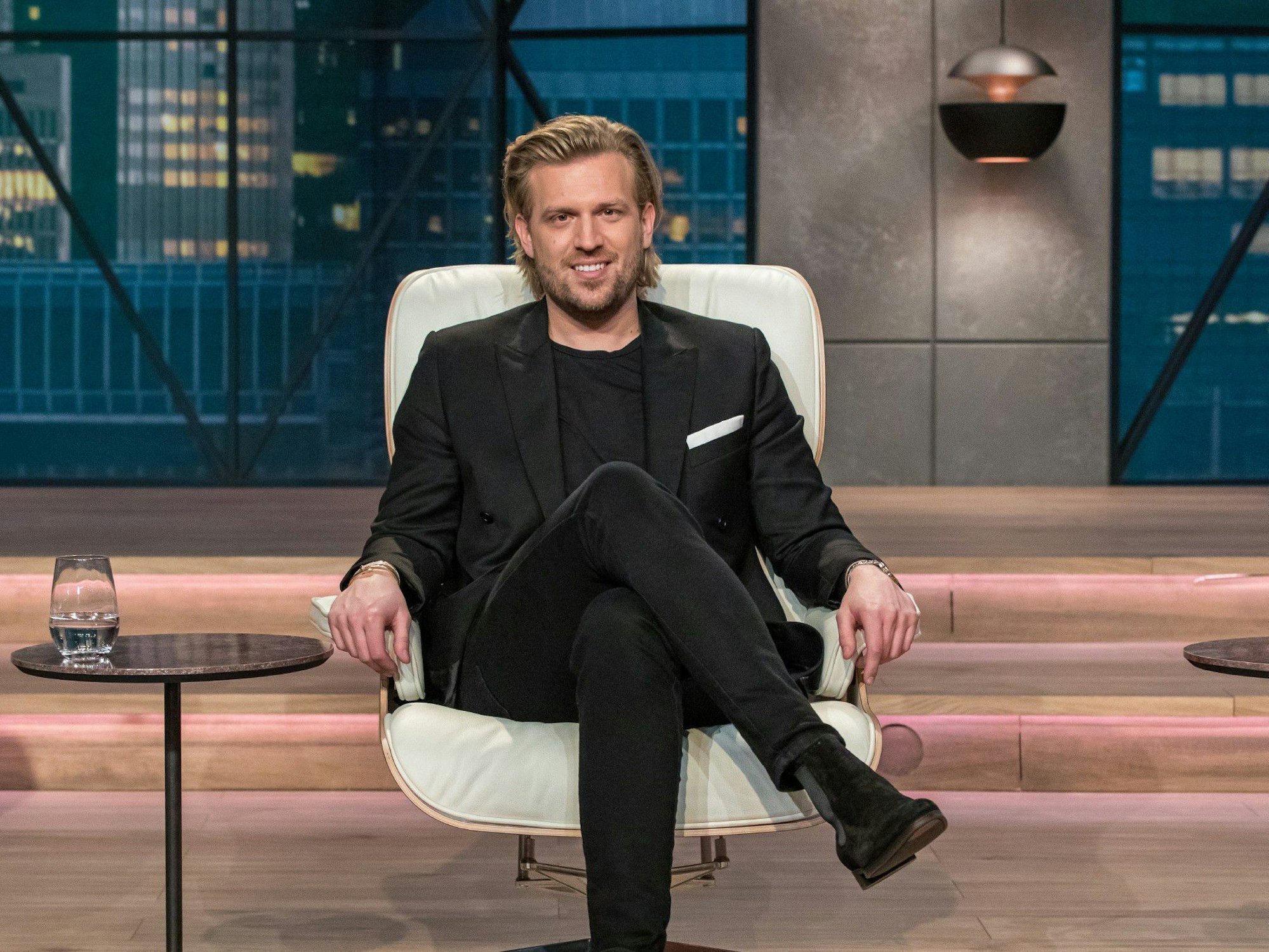 Investor Tillman Schulz sitzt auf einem Stuhl im Studio von Die Höhle der Löwen. Er trägt einen dunklen Anzug und ein dunkles T-Shirt. Die Verwendung des sendungsbezogenen Materials ist nur mit dem Hinweis und Verlinkung auf RTL+ gestattet.