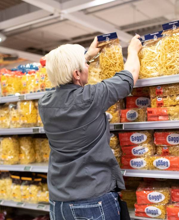 Das Symbolfoto aus dem Jahr 2020 zeigt eine Supermarkt-Mitarbeiterin mit grauem Hemd und kurzen blonden Haaren vor einem Regal mit allen möglichen Nudelsorten.