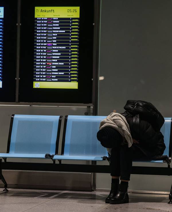 Eine Frau sitzt am Flughafen Köln/Bonn neben einer Anzeigetafel, auf der zahlreiche Flüge annulliert sind.