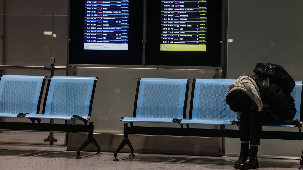 Eine Frau sitzt am Flughafen Köln/Bonn neben einer Anzeigetafel, auf der zahlreiche Flüge annulliert sind.