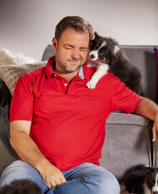 Hundeprofi Martin Rütter sitzt vor einer Couch und kuschelt mit einigen Hundewelpen.