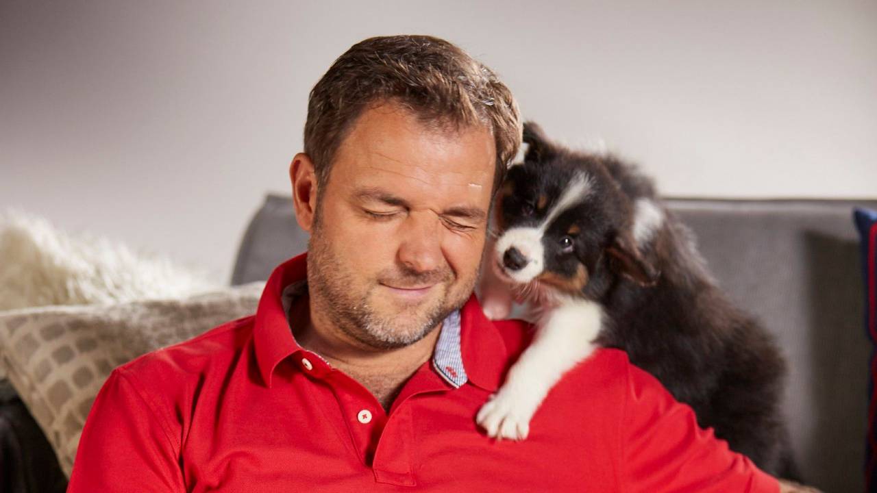 Hundeprofi Martin Rütter sitzt vor einer Couch und kuschelt mit einigen Hundewelpen.
