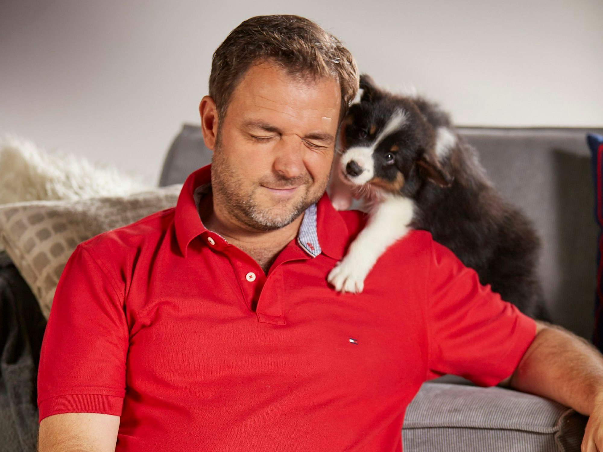 Hundeprofi Martin Rütter sitzt vor einer Couch und kuschelt mit einigen Hundewelpen.