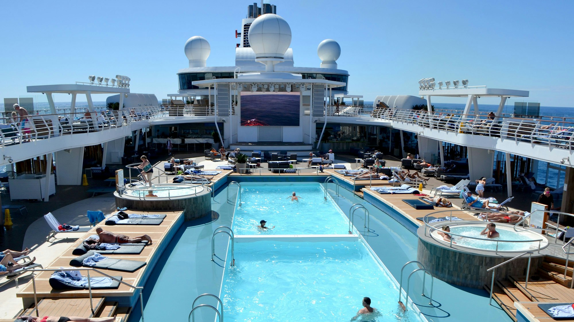 Ein Bild des Pools an Deck des Schiffes. Auf den Liegen rund um den Pool liegen einige Menschen und sonnen sich.