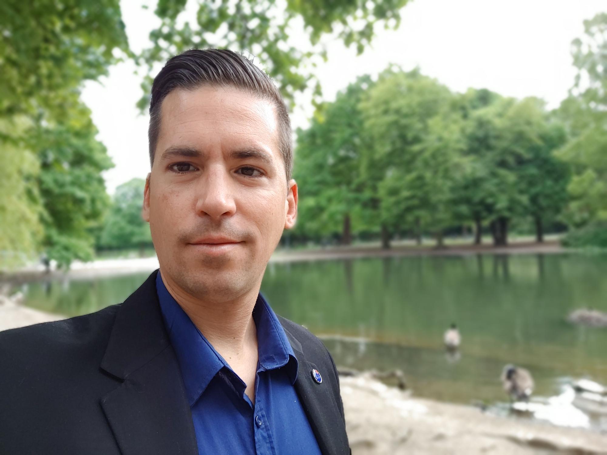 Tolga Özgül steht vor einem See und blickt für ein Selfie in die Kamera.