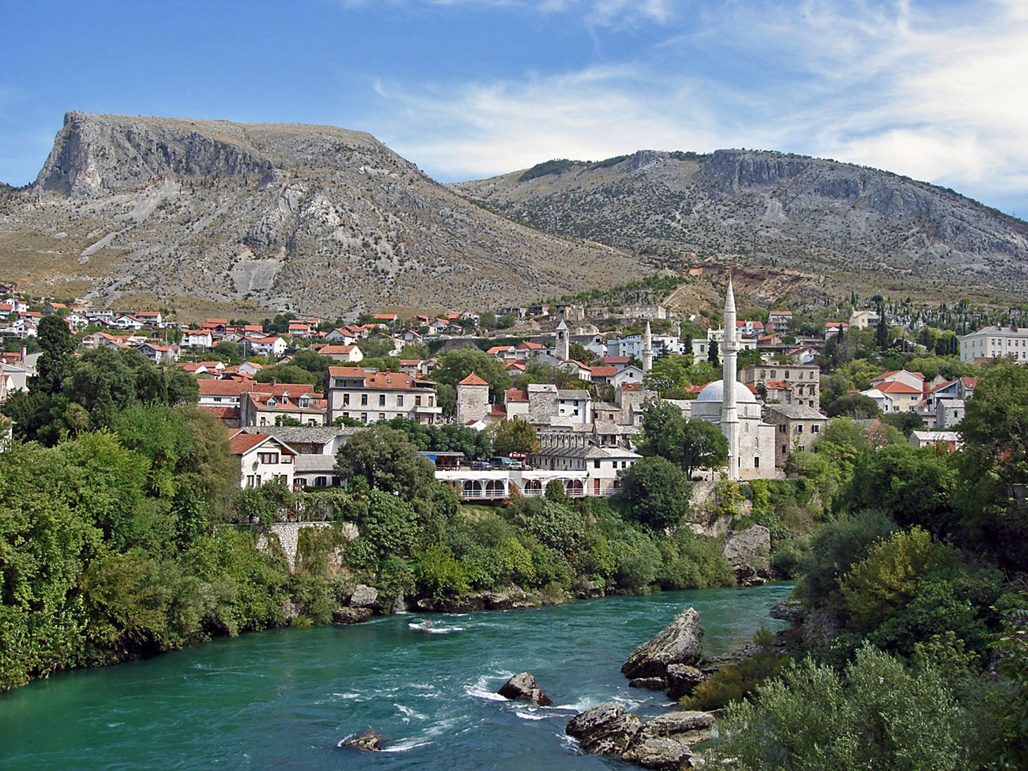 Das Foto zeigt einen schmalen Fluss, der an einem kleinen Ort in Bosnien-Herzegowina vorbeifließt. Im Hintergrund sind Berge zu sehen.