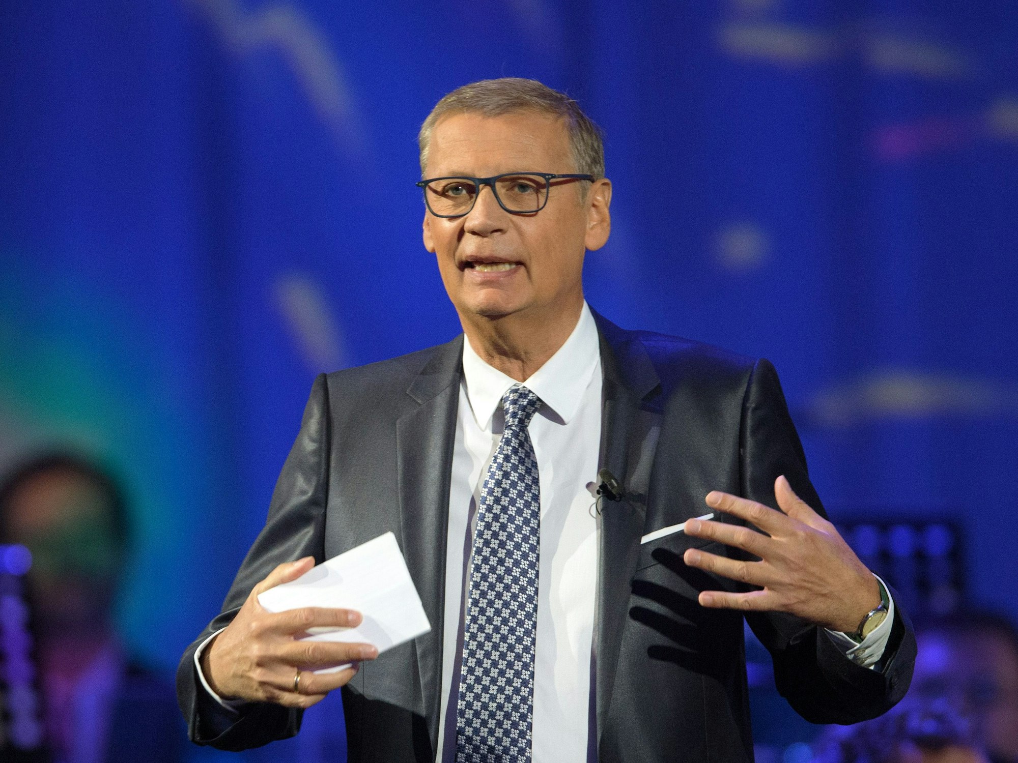 Günther Jauch gestikuliert und spricht. Er trägt einen Anzug und hält eine Moderationskarte in der Hand.