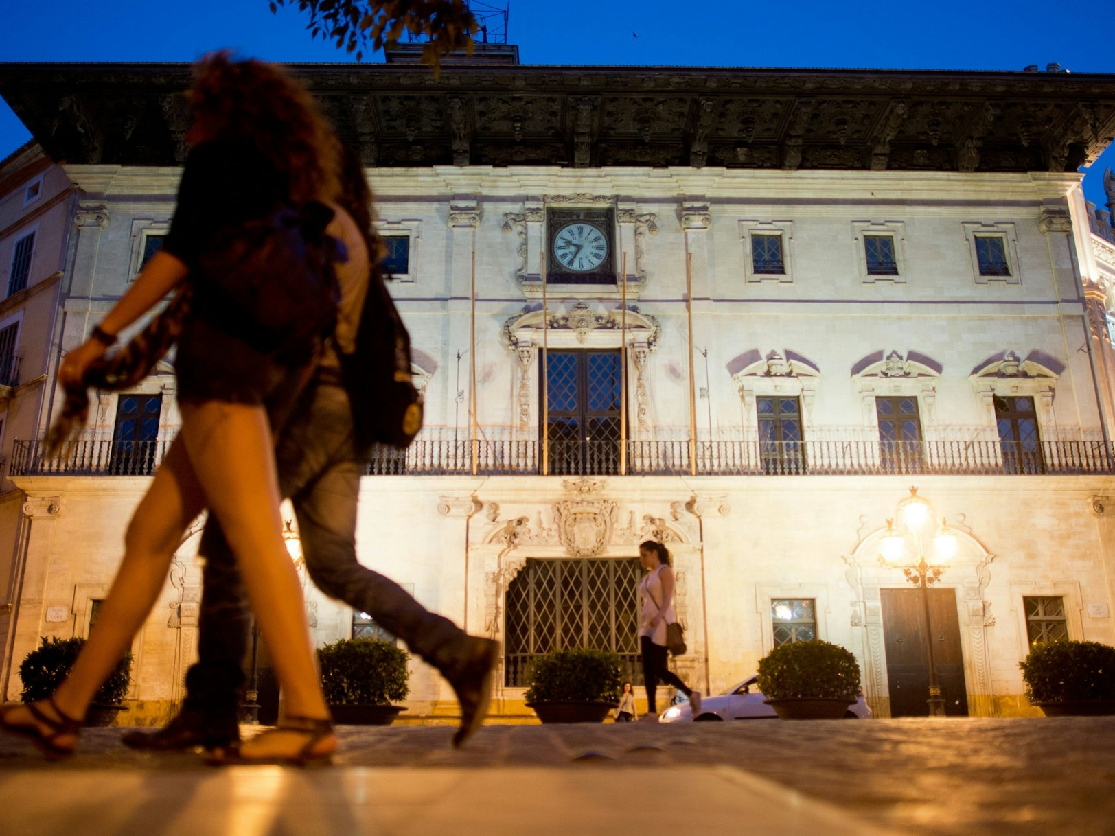 Das Bild zeigt das Rathaus in Palma de Mallorca bei Nacht. Zwei Passanten laufen im Vordergrund vorbei.