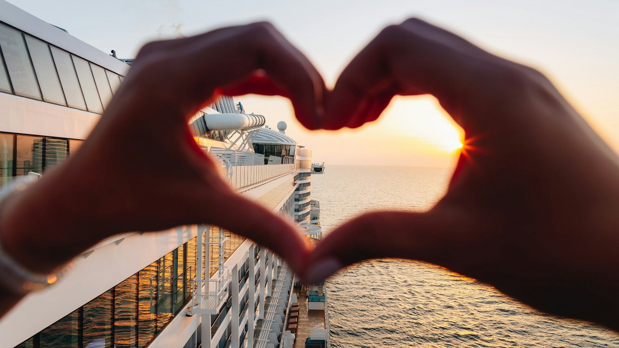 Ein Urlauber steht an Deck eines Schiffes und formt mit den Händen ein Herz. Im Hintergrund geht die Sonne im Meer unter.
