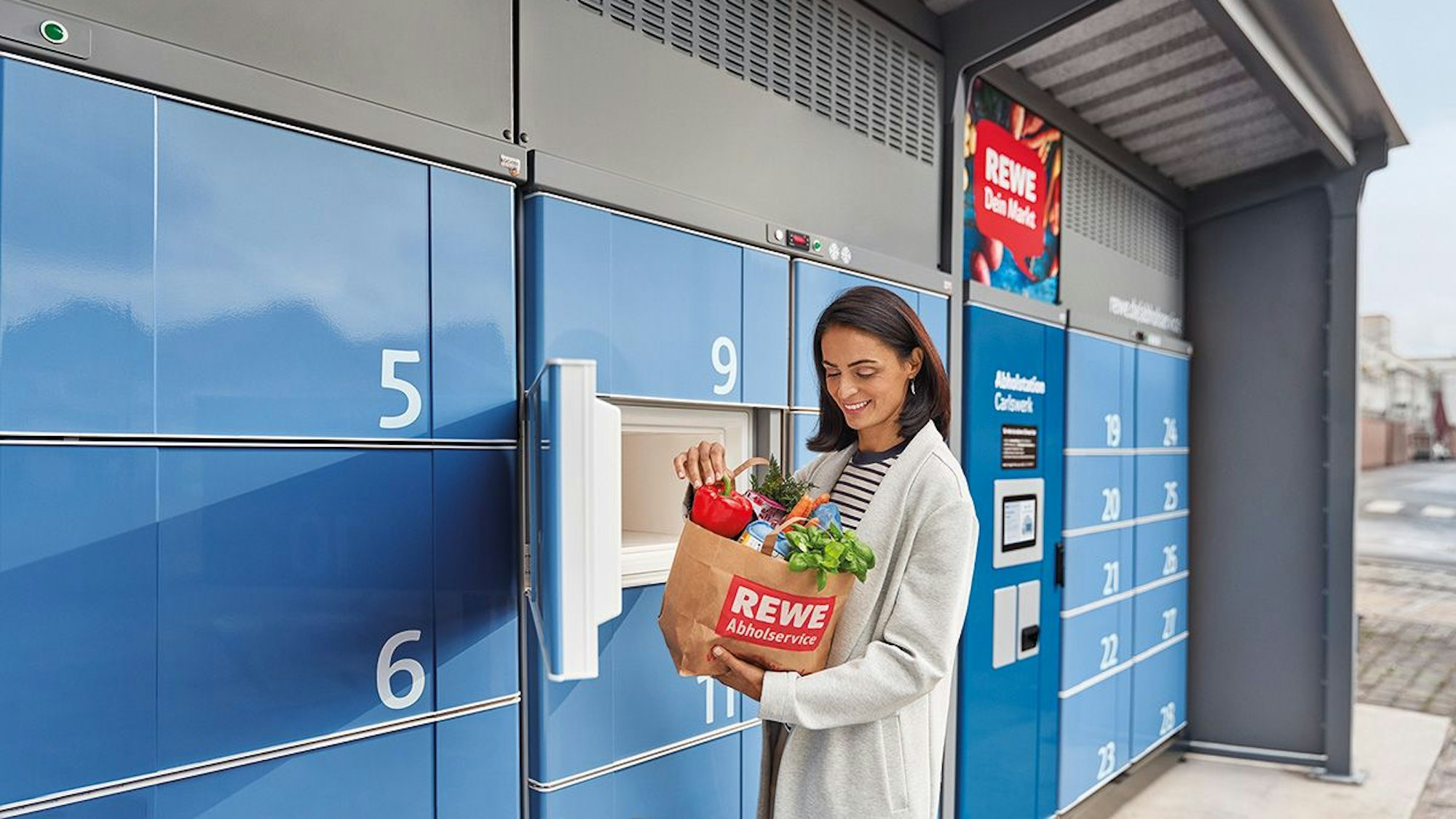 Eine Frau steht vor einer Abholstation von Rewe.