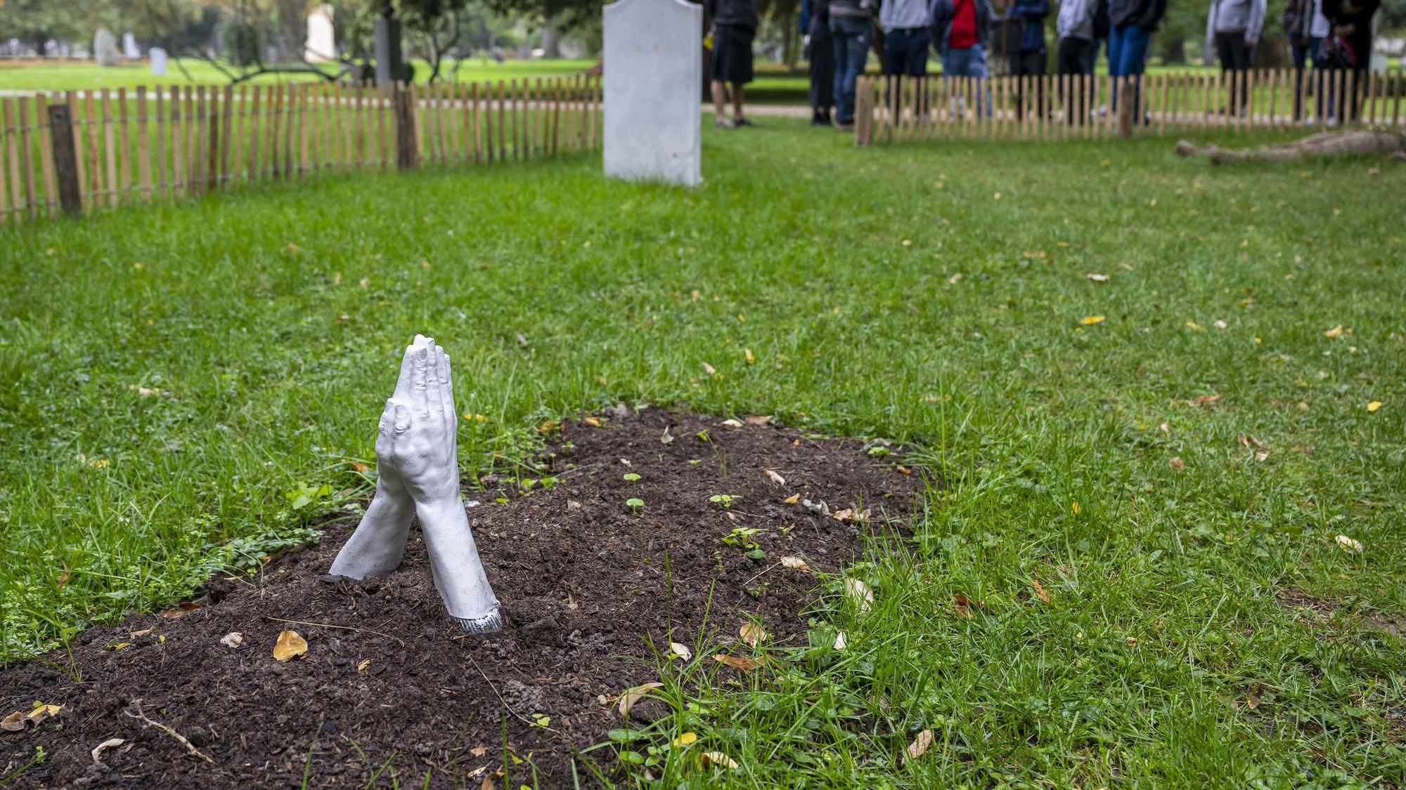 Das Symbolfoto aus dem Jahr 2022 zeigt ein frisches Grab auf einer grünen Wiese. Aus der Erde ragen zwei steinerne Hände, die zum Gebet aneinandergelehnt sind.