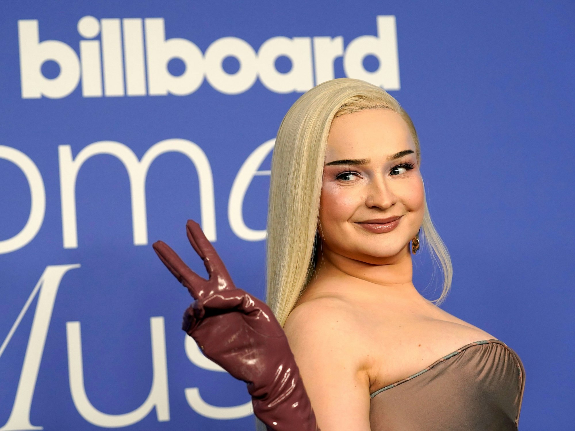 Das Foto aus dem Jahr 2023 zeigt die deutsche Sängerin Kim Petras. Die Blondine posiert vor einer blauen Wand, trägt ein braunes Kleid und Lack-Handschuhe.