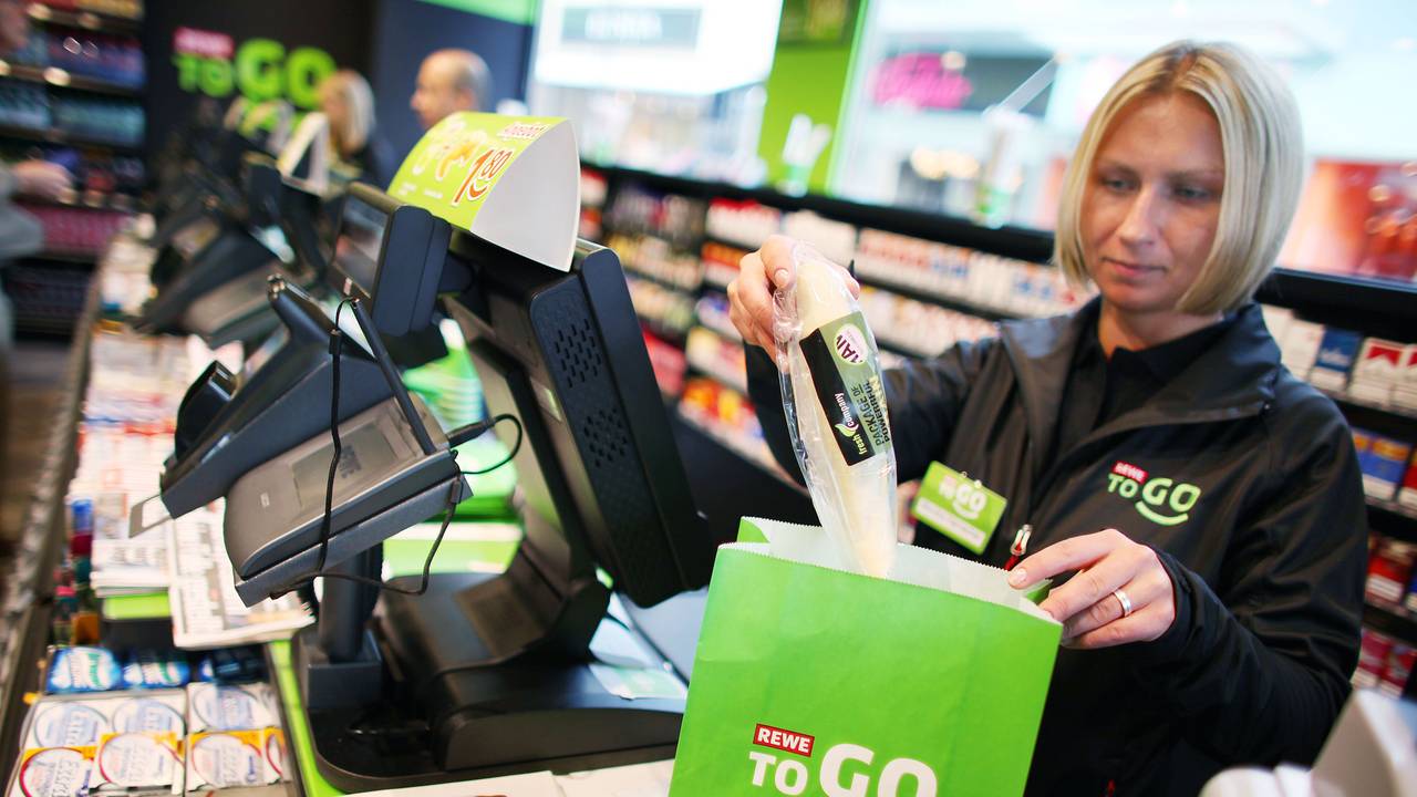 Das Symbolfoto aus dem Jahr 2011 zeigt eine blonde Kassiererin einer Rewe-To-Go-Filiale. Die Frau steht hinter der Kasse und packt Produkte in eine grüne Papiertüte.