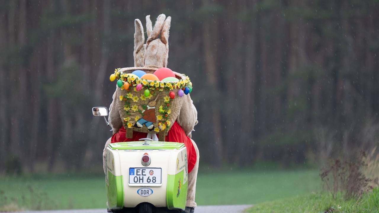 Das Symbolfoto aus dem Jahr 2023 zeigt einen alten Roller, auf dem zwei Personen in Hasenkostümen sitzen. Hinten auf dem Roller befindet sich ein Korb mit bunten Ostereiern.