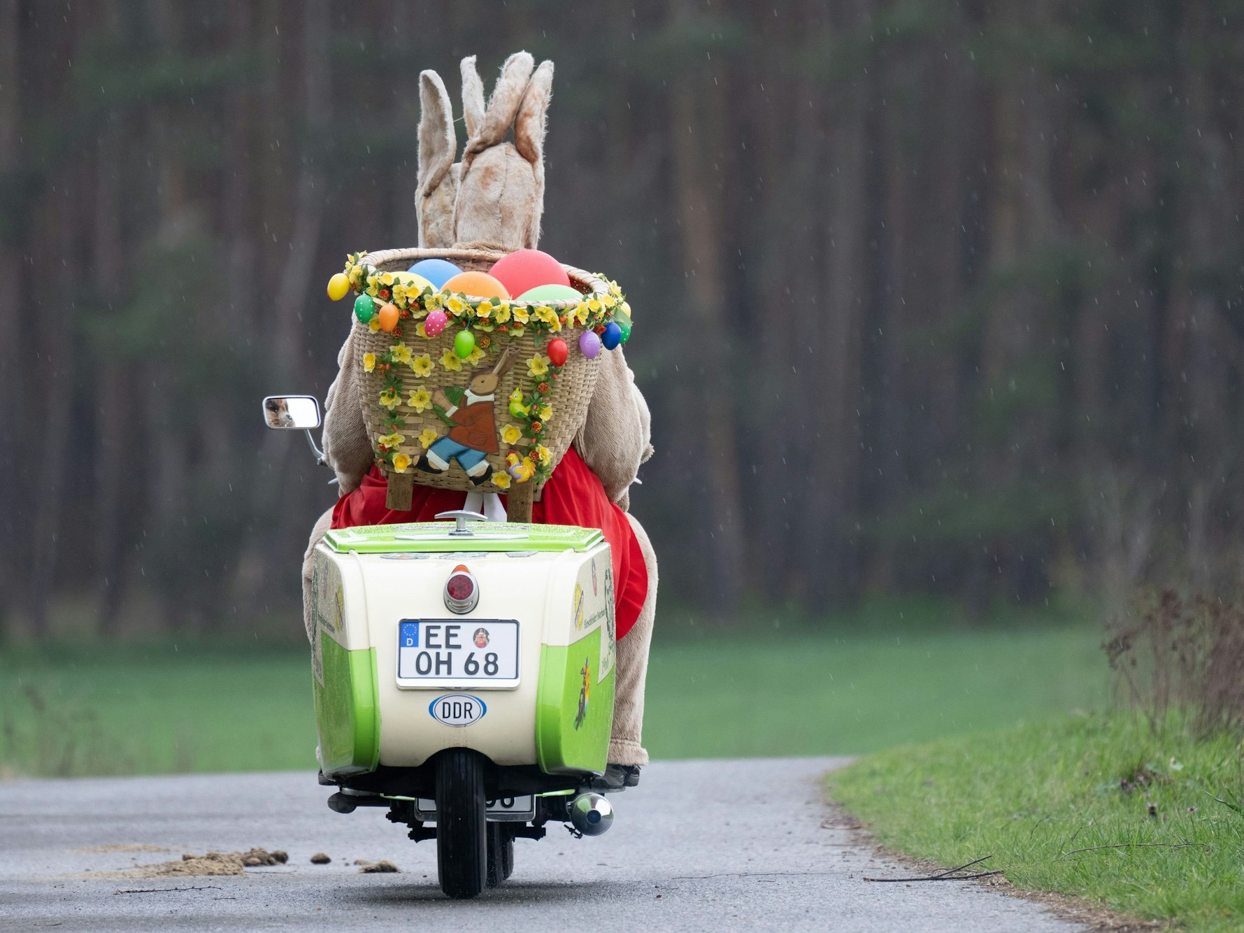 Das Symbolfoto aus dem Jahr 2023 zeigt einen alten Roller, auf dem zwei Personen in Hasenkostümen sitzen. Hinten auf dem Roller befindet sich ein Korb mit bunten Ostereiern.