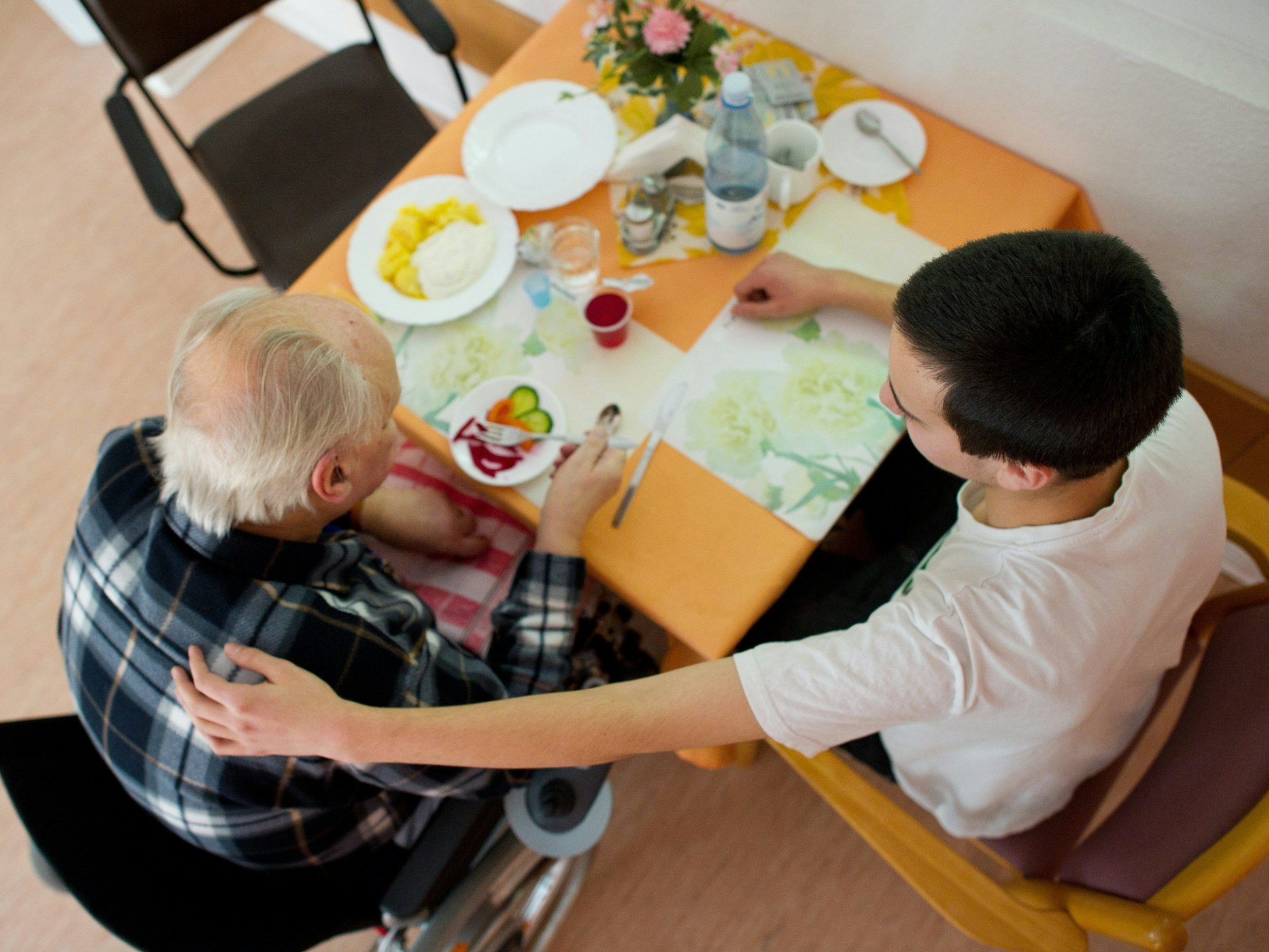Auf dem Archivbild sieht man einen Mann im Rollstuhl am Esstisch. Neben ihm sitzt ein Pfleger, der seinen Arm um den Mann legt.