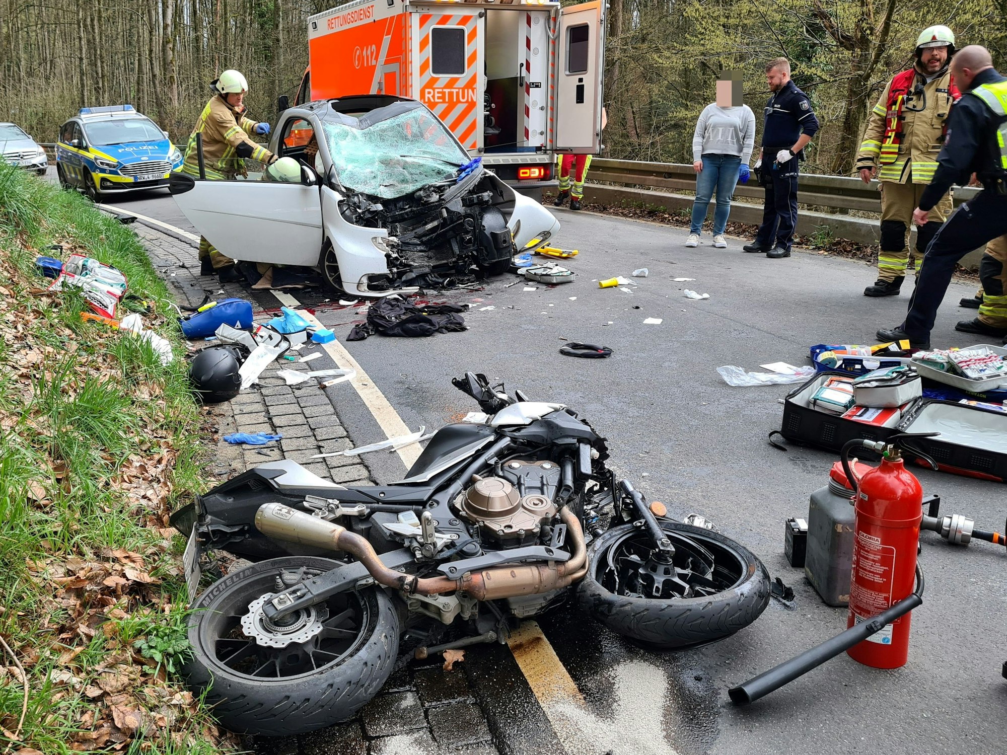 Nach einem Unfall steht ein Smart mit zerborstener Windschutzscheibe und zerfetztem Motorraum auf der Fahrbahn, ein paar Meter davor liegt ein Motorrad. Dazwischen Trümmerteile und persönliche Gegenstände.