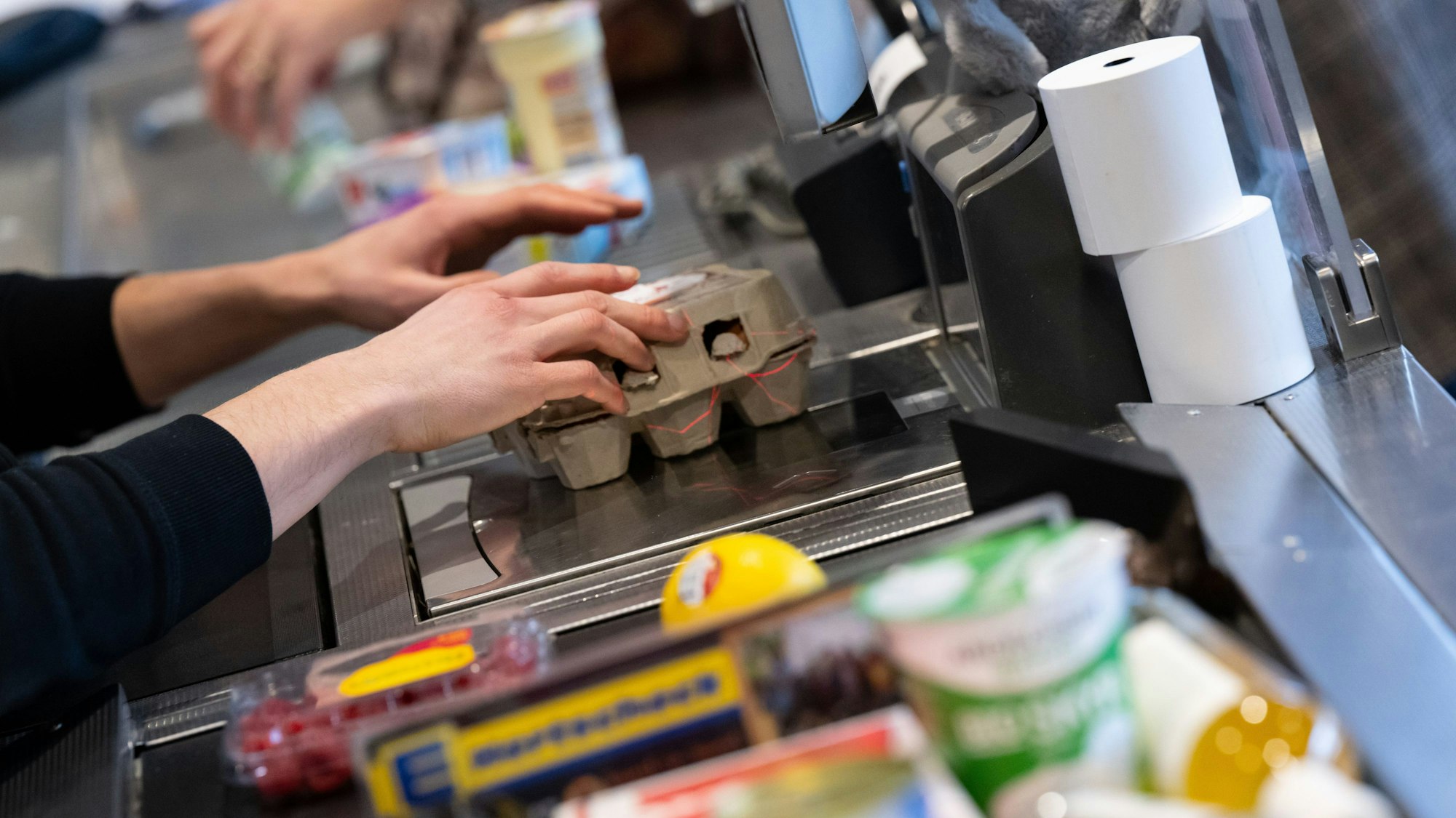 Das Symbolfoto aus dem Jahr 2023 zeigt ein Kassenband mit Lebensmitteln. Eine Kassiererin zieht eine Packung Eier über den Scanner.