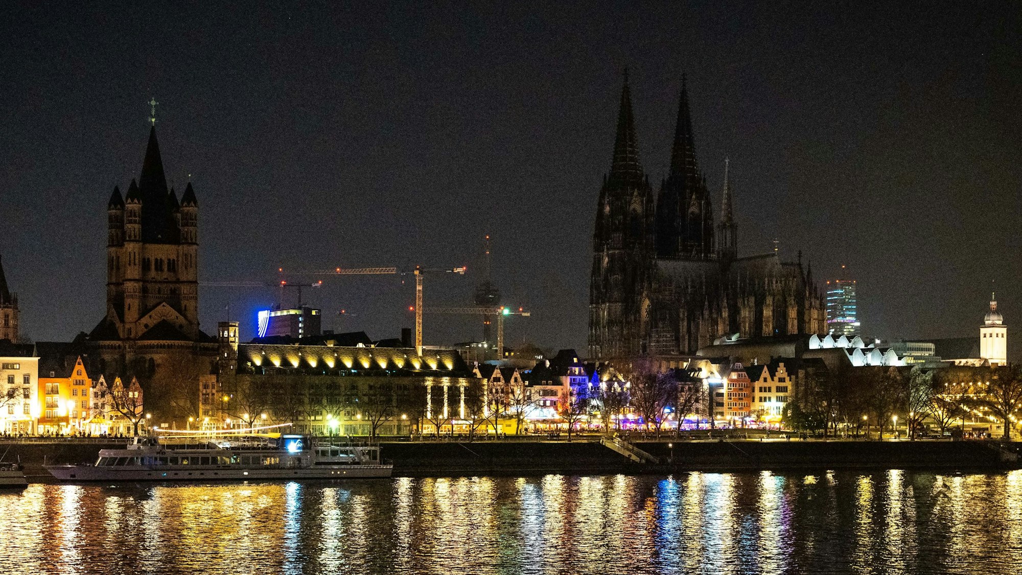 Die beleuchtete Rheinpromenade in Köln in der Dunkelheit. Im Hintergrund ist der Kölner Dom zu sehen, der nicht beleuchtet ist.