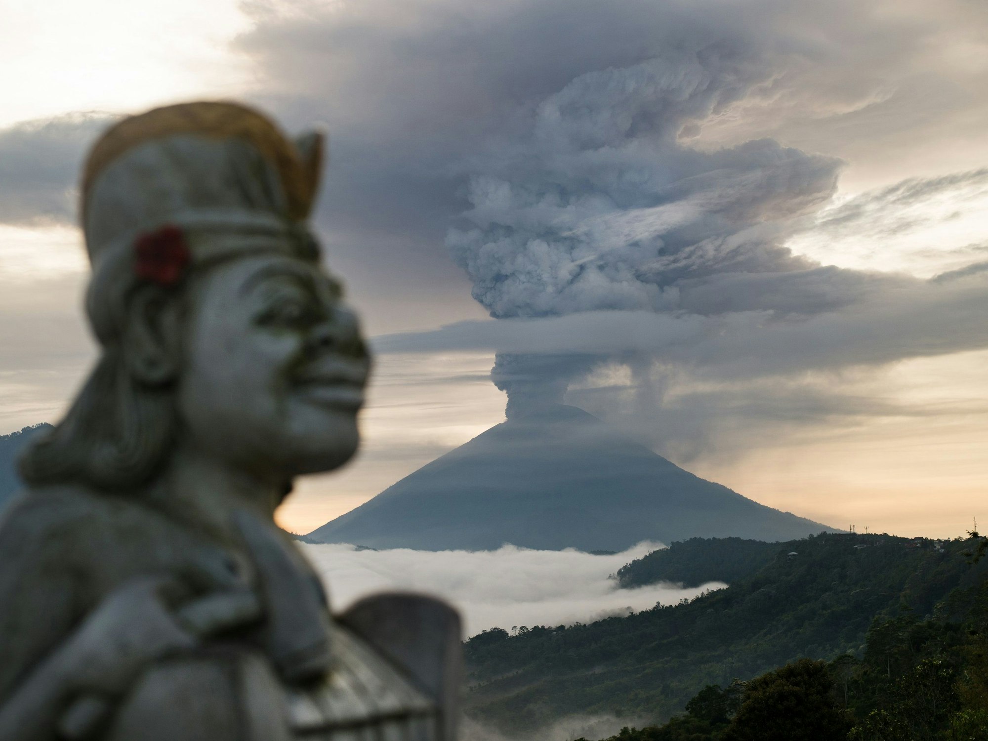Der heilige Berg Agung auf der indonesischen Insel Bali während eines Vulkanausbruches. Rauch steigt aus dem Vulkan empor. Im Vordergrund ist eine Statue zu sehen.