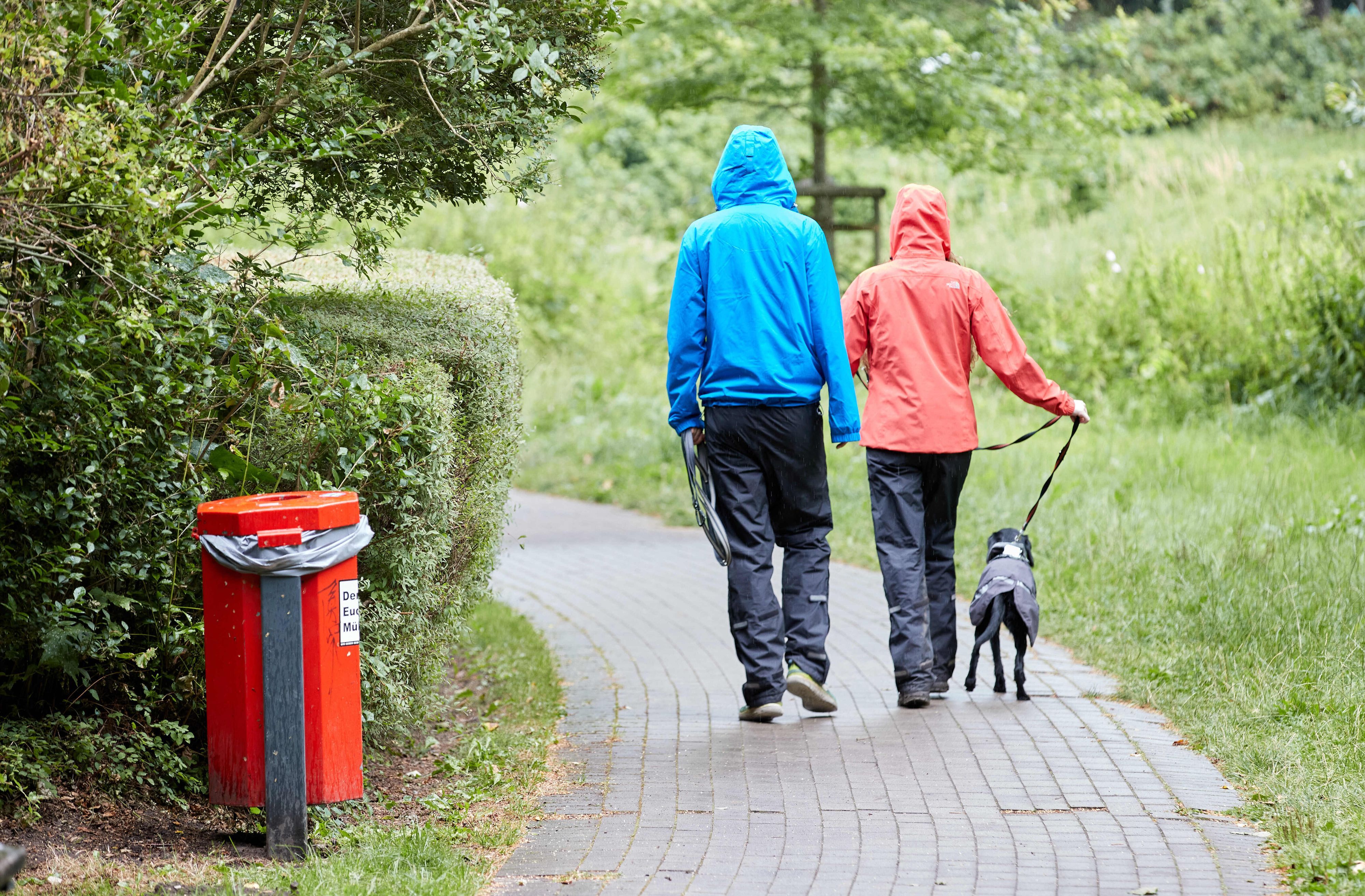 Ein Mann, eine Frau und ein Hund gehen mit Regenschutzkleidung in einer Grünfläche in Hamburg-Lokstedt spazieren.