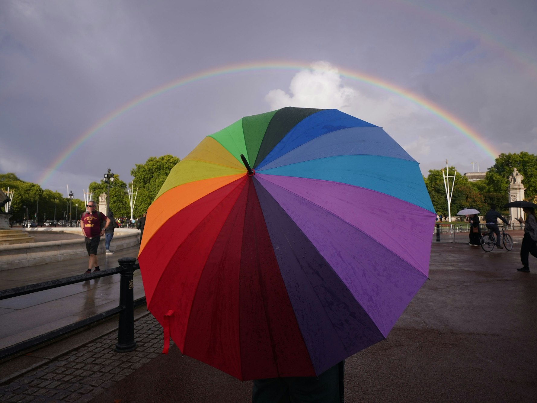 Ein Regenbogen ist zu sehen, während sich eine Person mit einem Regenschirm in Regenbogenfarben im September 2022 vor dem Buckingham Palace befindet.