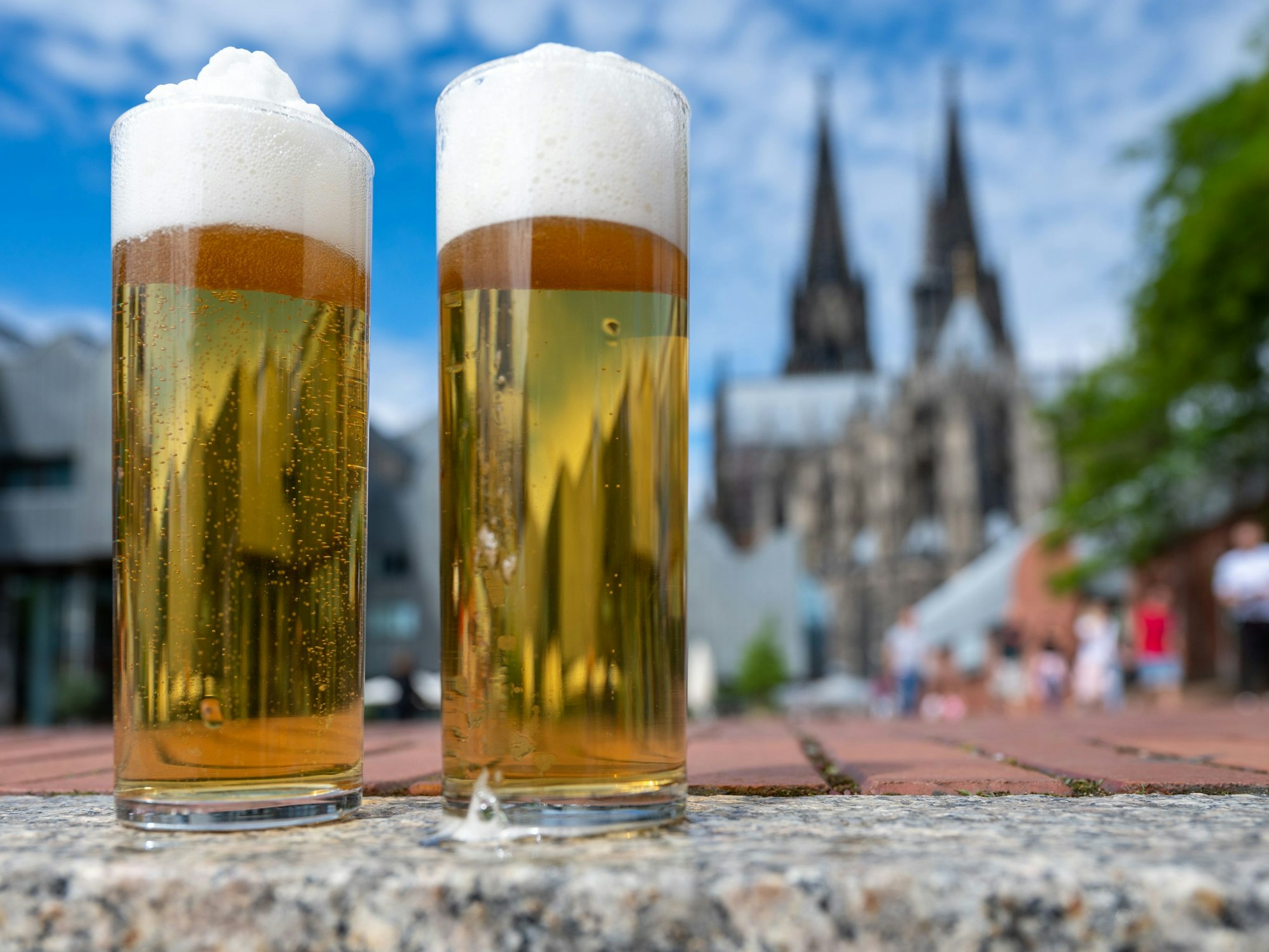 Zwei Kölsch-Gläser stehen auf einer Mauer, im Hintergrund ist der Dom zu sehen.