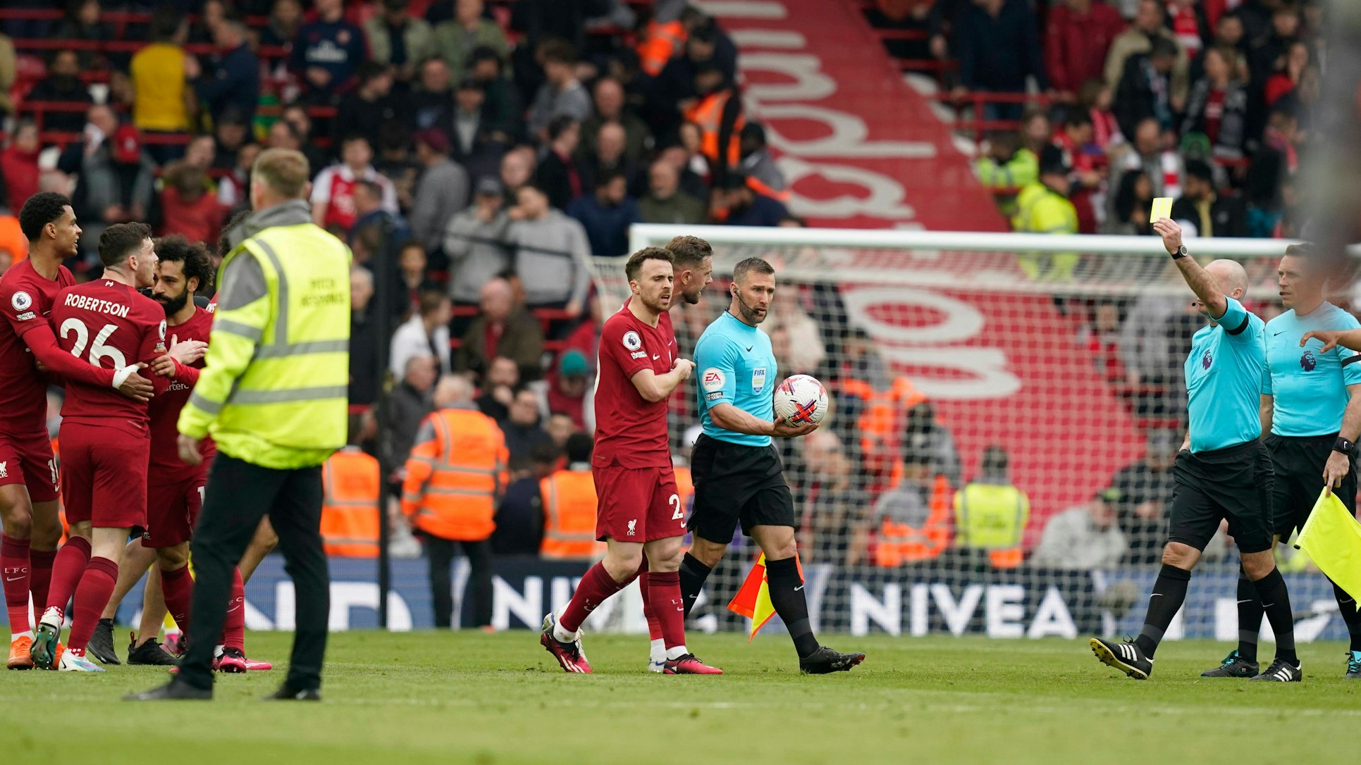 Referee Paul Tierney (r.) zeigt Andrew Robertson (l.) vom FC Liverpool die Gelbe Karte. Zuvor hatte Linienrichter Constantine Hatzidakis (M.) ihn per Ellbogenstoß attackiert.