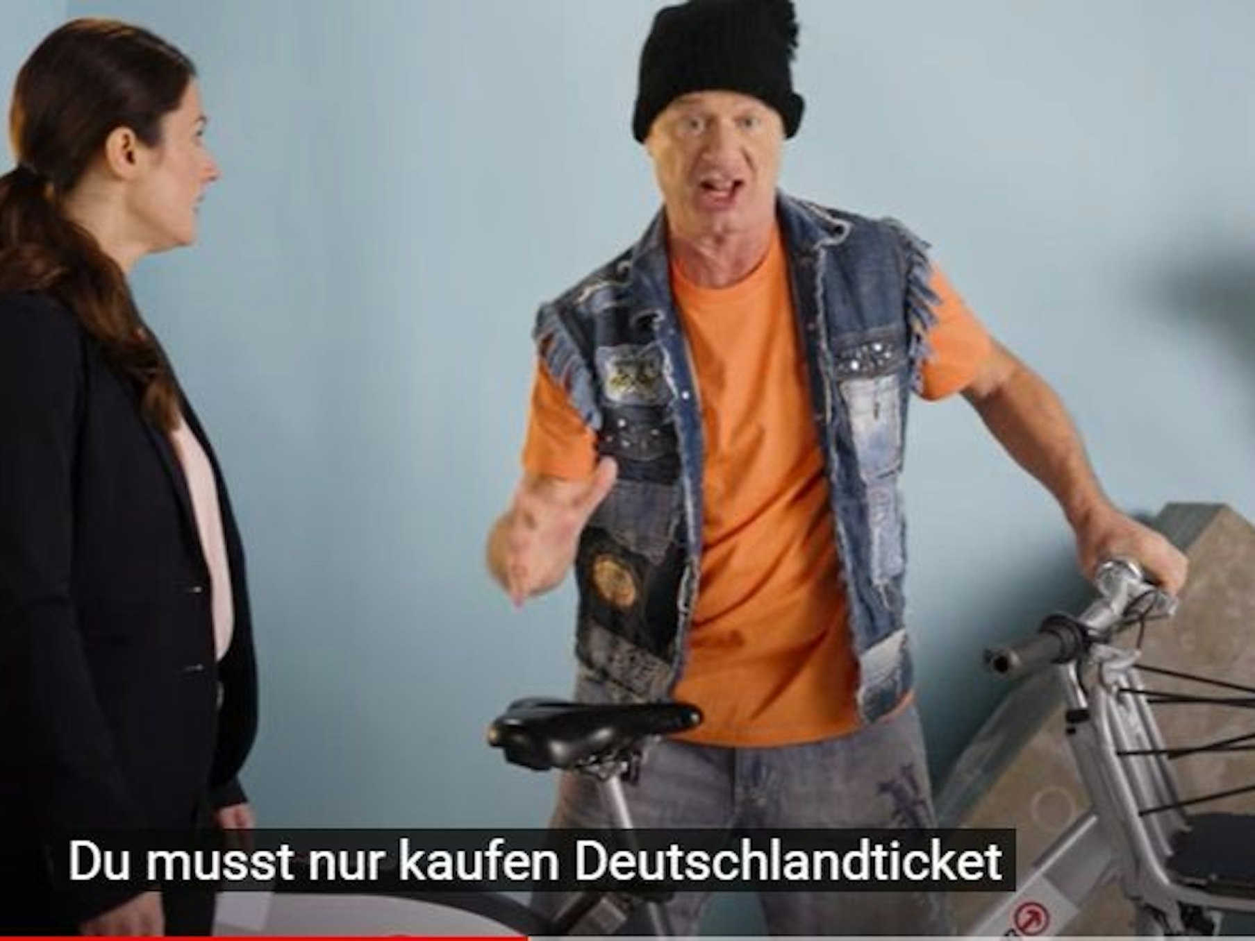 Tom Gerhardt, mit Bommelmütze auf dem Kopf, steht neben einem Fahrrad der KVB und macht Werbung für das Deutschlandticket.