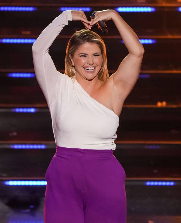 Beatrice Egli steht auf der Bühne mit einem weißen Oberteil und einer lila Hose und formt mit ihrem Armen ein Herz.