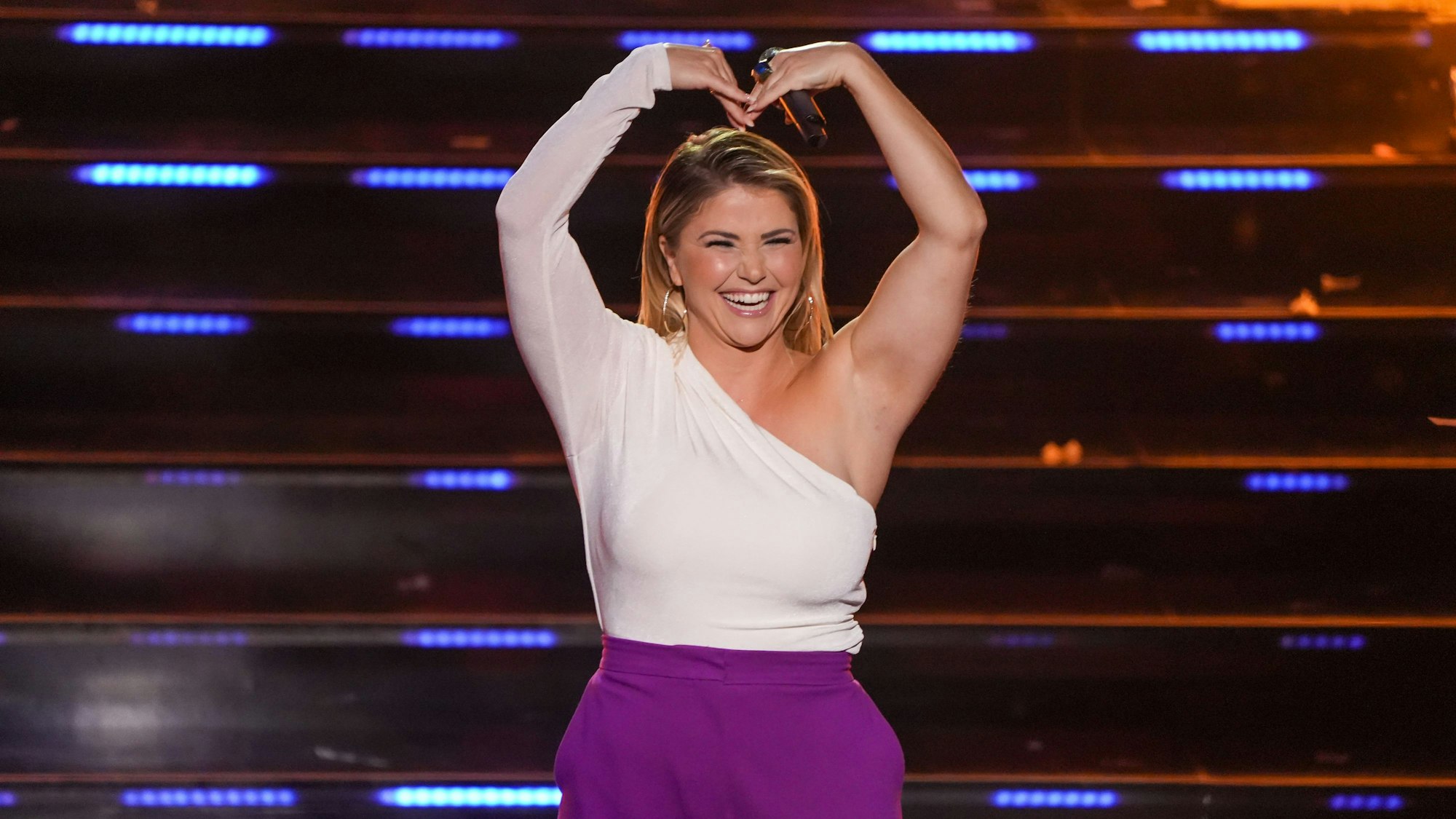 Beatrice Egli steht auf der Bühne mit einem weißen Oberteil und einer lila Hose und formt mit ihrem Armen ein Herz.