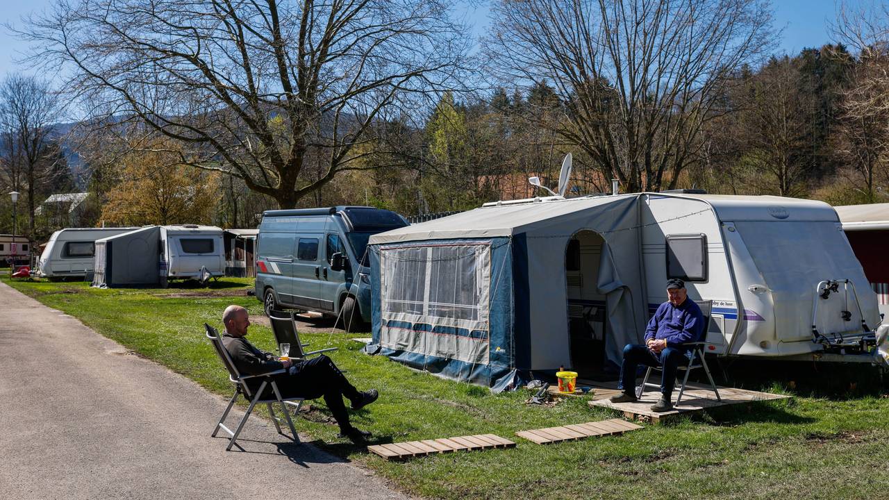 Zwei Männer sitzen in der Sonne vor einem Wohnwagen auf einem Campingplatz.
