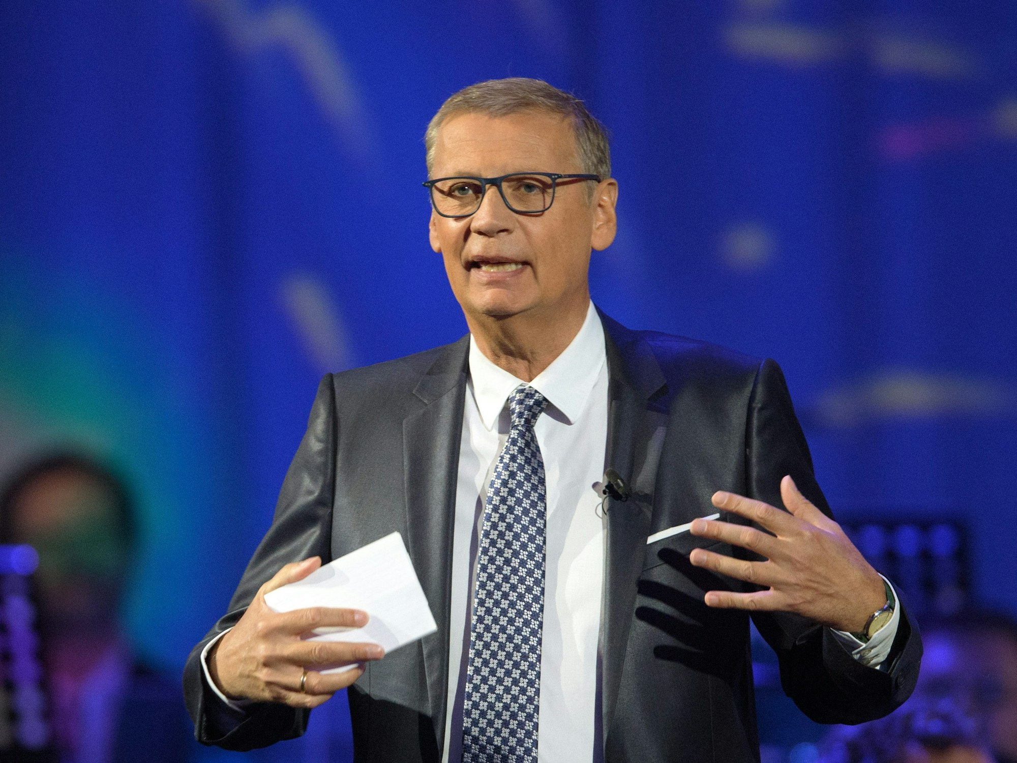 Günther Jauch moderiert im Anzug, hier im Oktober 2020, mit karierter Krawatte eine Talkrunde und hält dabei Karten in der Hand.