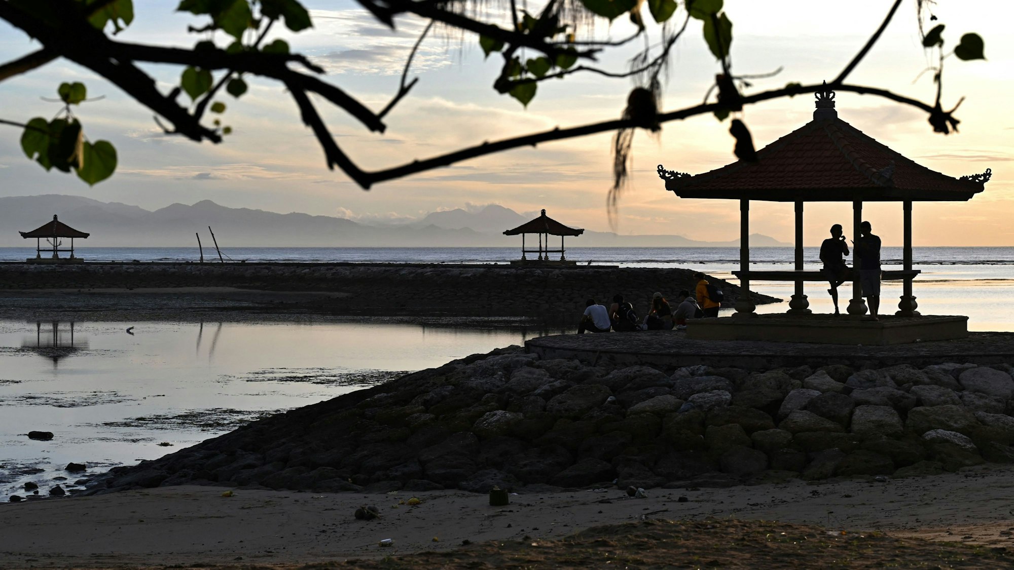 Das Foto aus dem Jahr 2023 zeigt einen Strandabschnitt auf der indonesischen Insel Bali. Auf Steinhügeln stehen einige Pavillons, um die sich Menschen scharen.