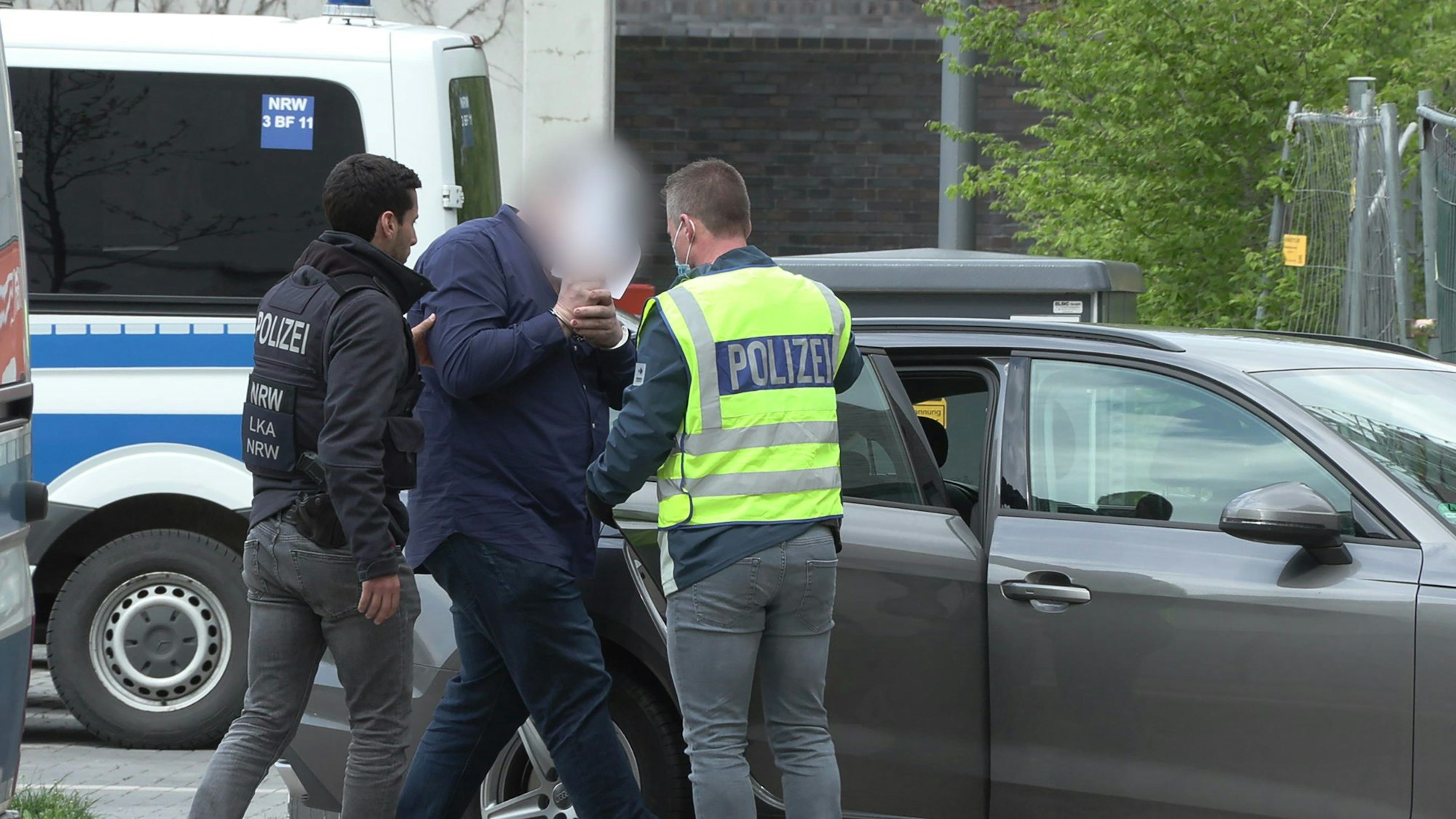 Zwei Männer, die Westen mit der Aufschrift „Polizei“ tragen, führen einen Mann in Handschellen zu einem Auto.