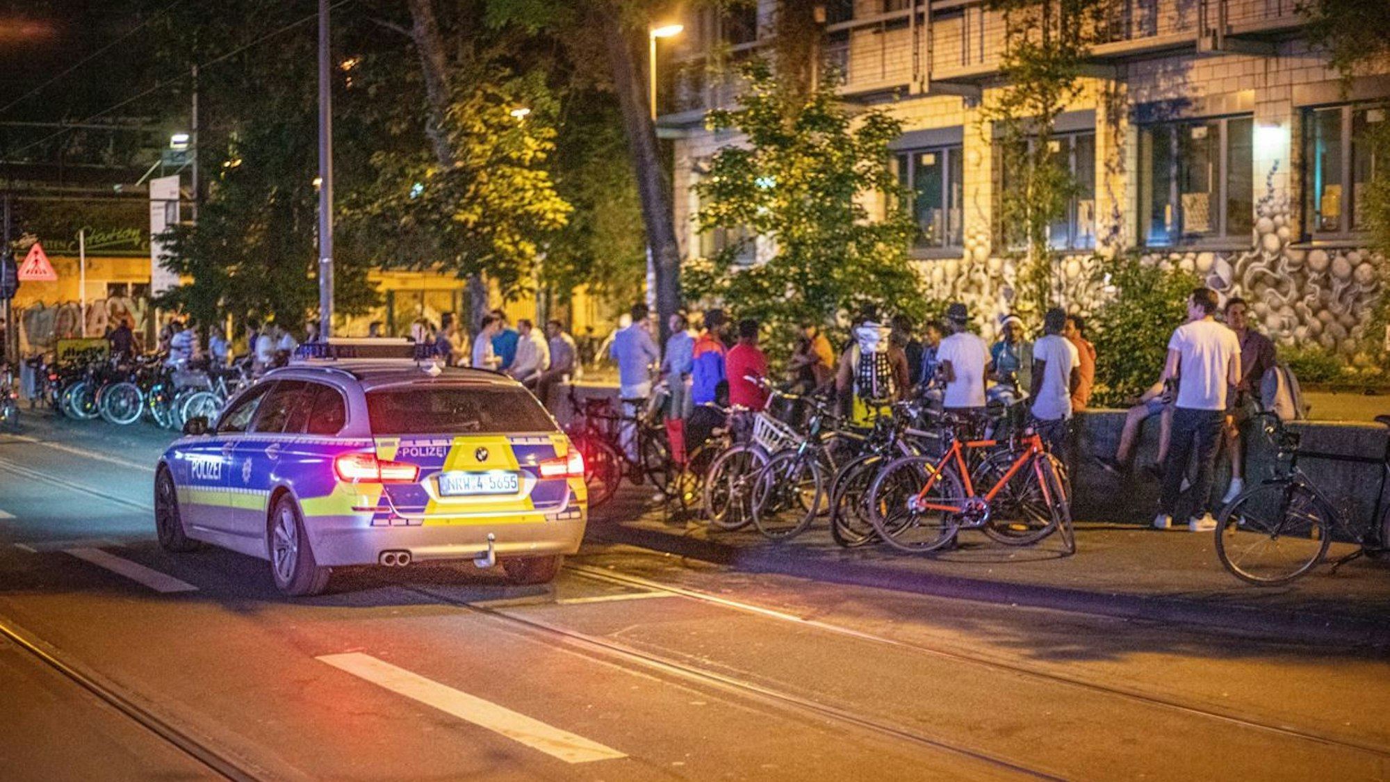 Menschen stehen an der Mauer an der Zülpicher Straße in Köln, daneben parkt ein Streifenwagen der Polizei.